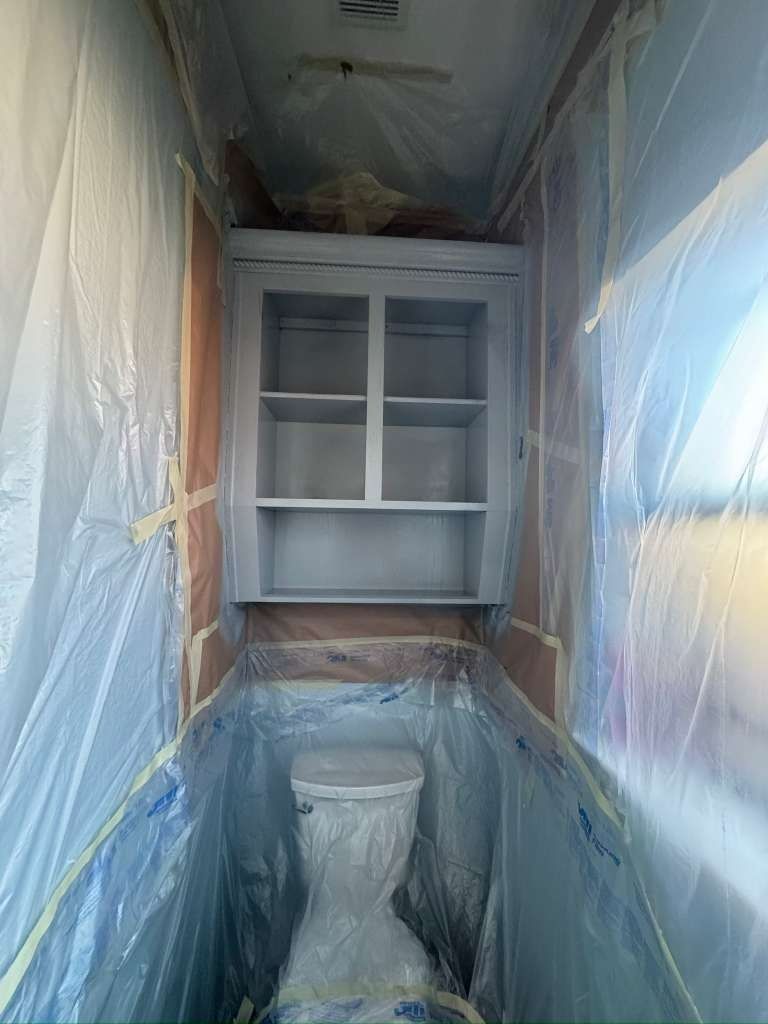 Narrow bathroom under renovation, wrapped in plastic with a toilet and wall cabinet visible.