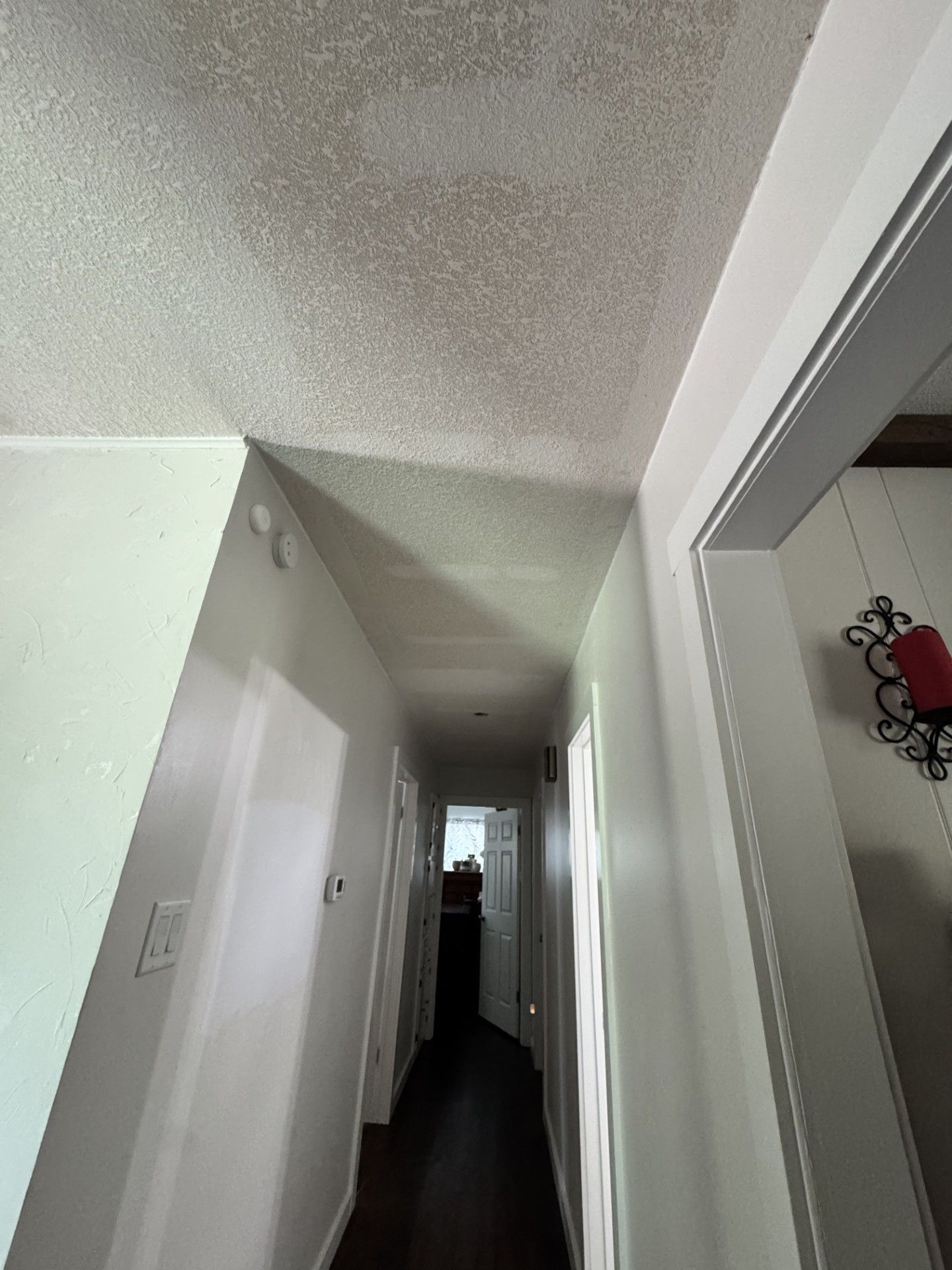 Narrow hallway with white walls, textured ceiling, and dark floor leading to a bright doorway.