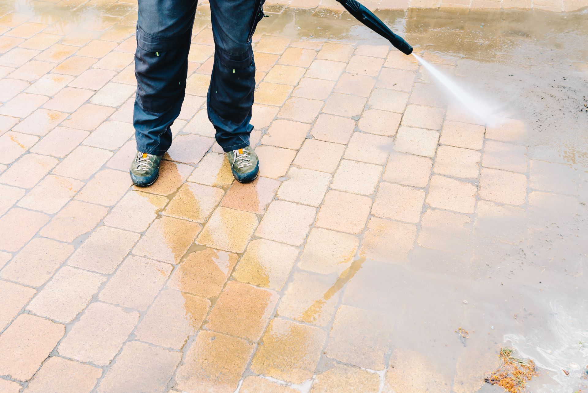 Person pressure-washing wet brick pavement with a hose, wearing jeans and sneakers.
