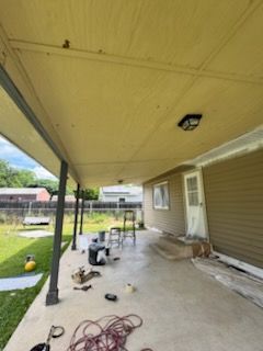 Covered backyard patio with tools and cords on the concrete floor beside a beige house
