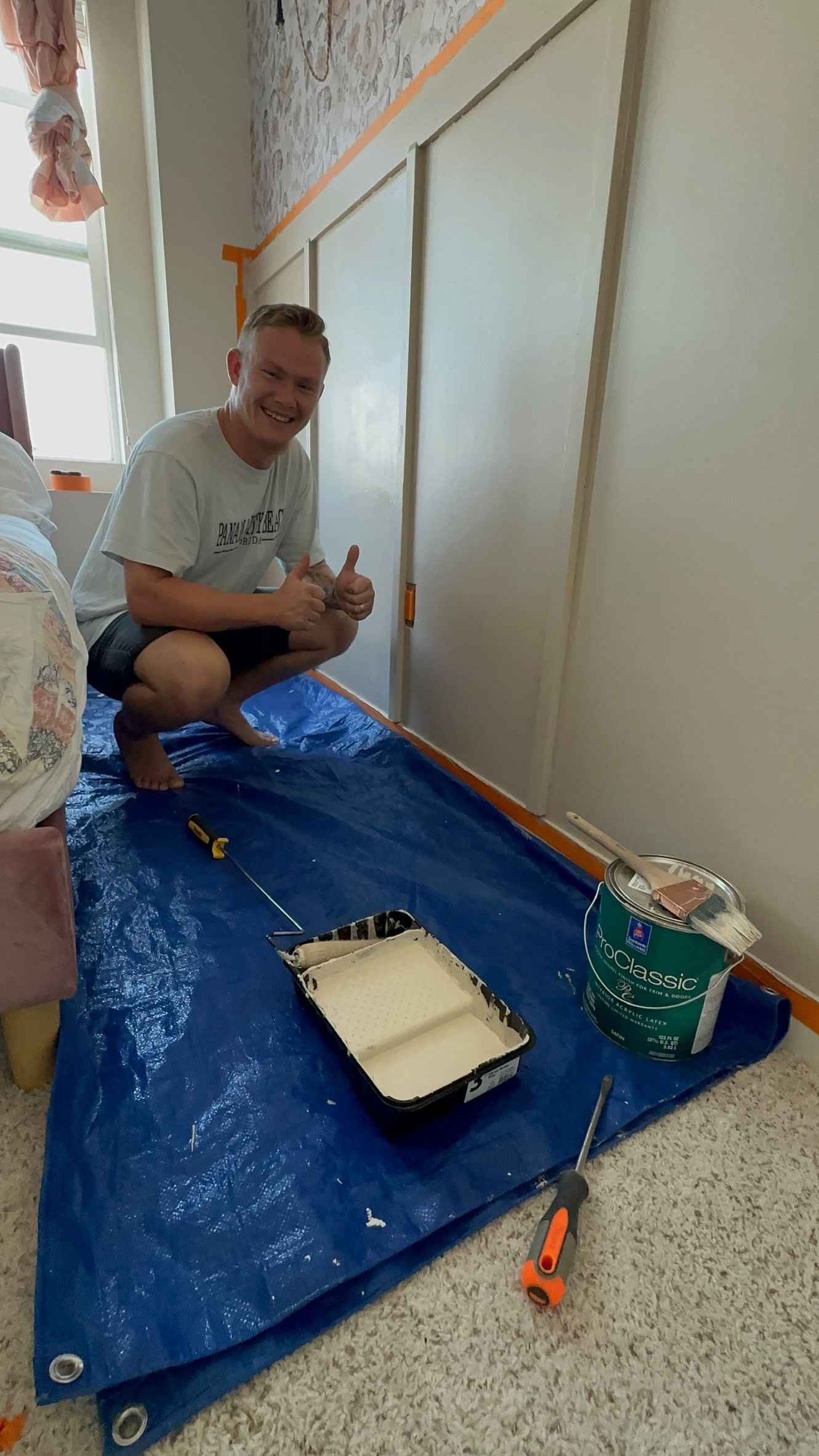 Person kneeling on a blue tarp, painting an interior wall with a tray and roller covered in plastic.