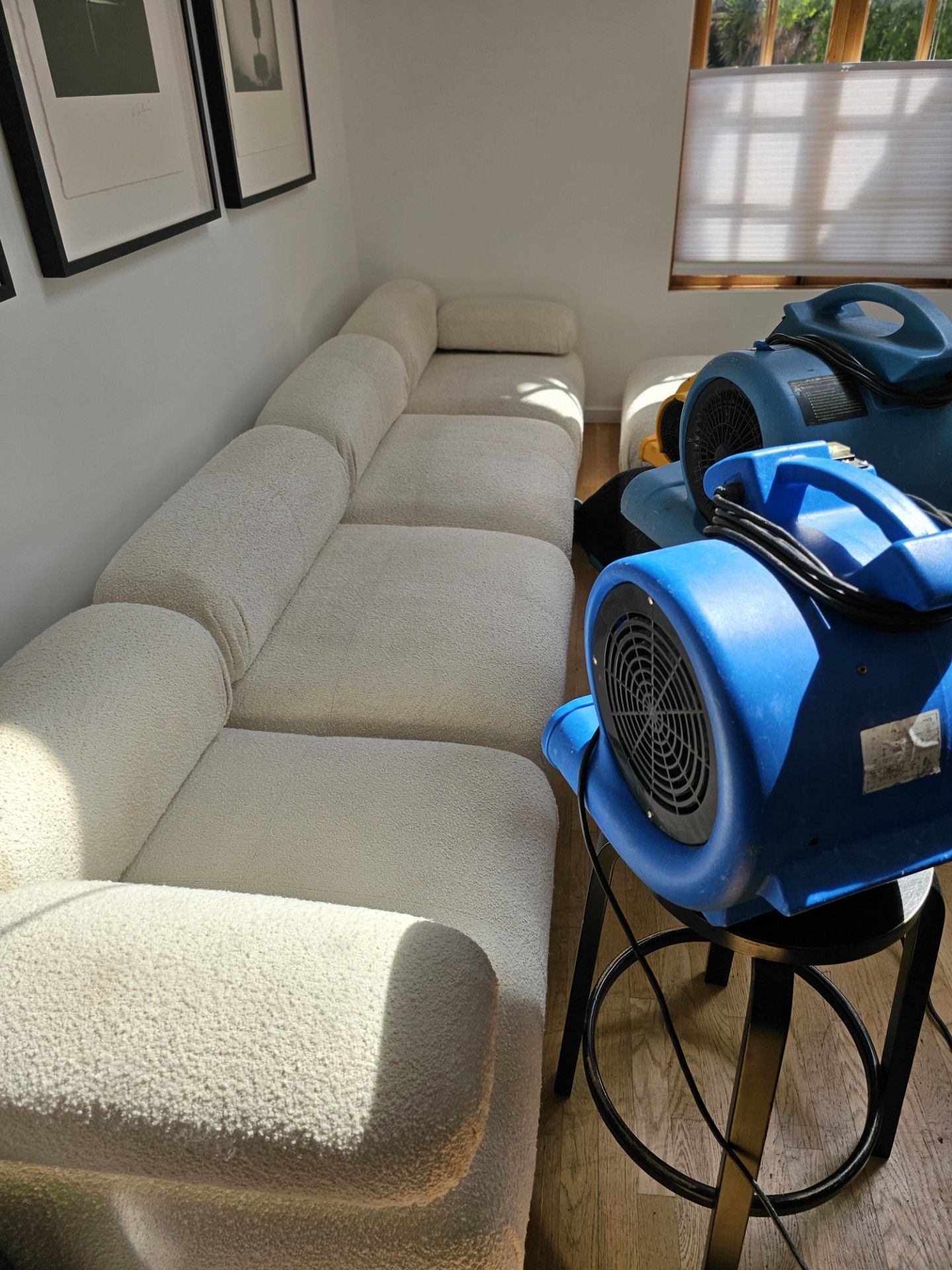 Small living room with beige sofa, wall art, window blinds, and two blue fans in front.