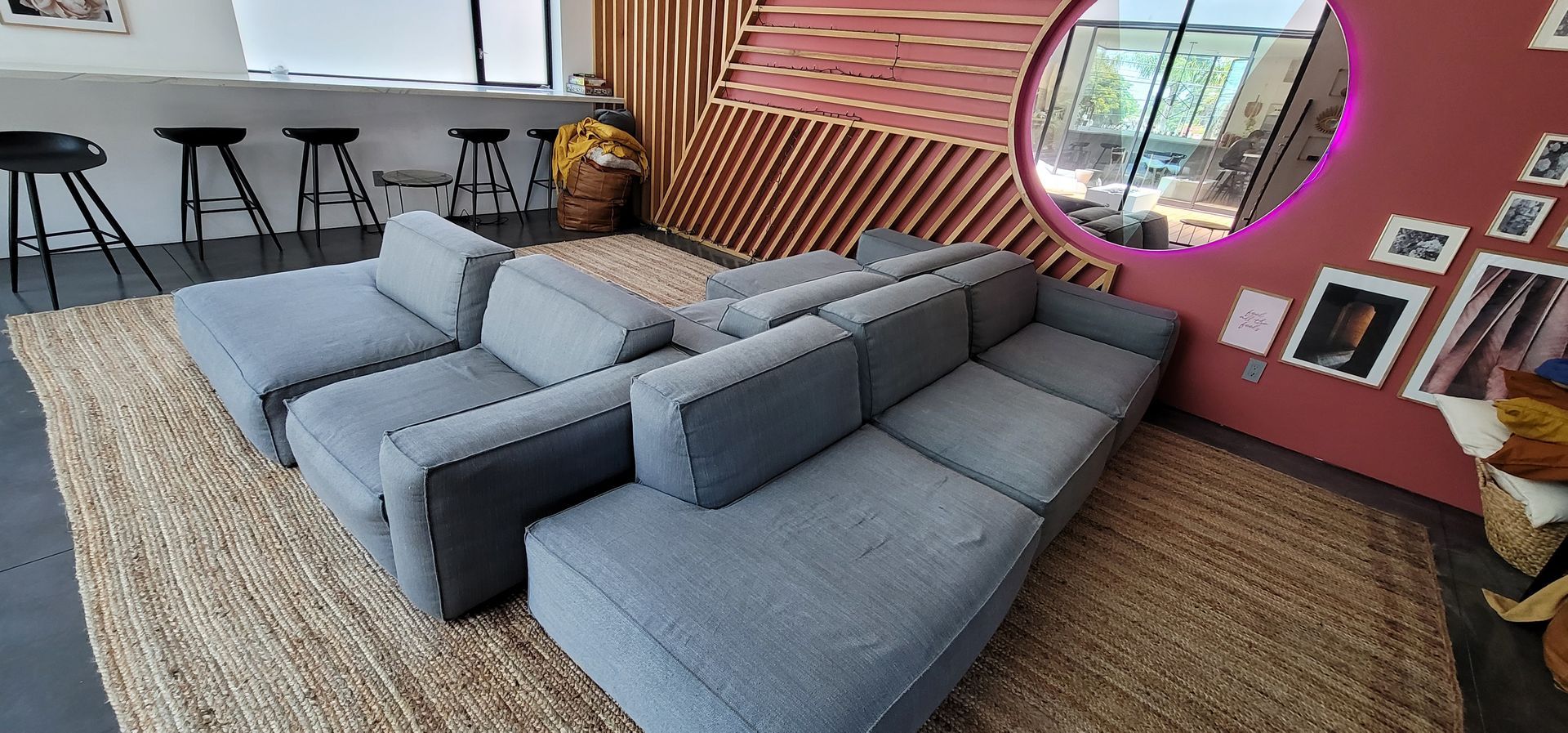 Modern lounge with gray sectional sofas, bar stools, woven rug, and a round window on a red wall.