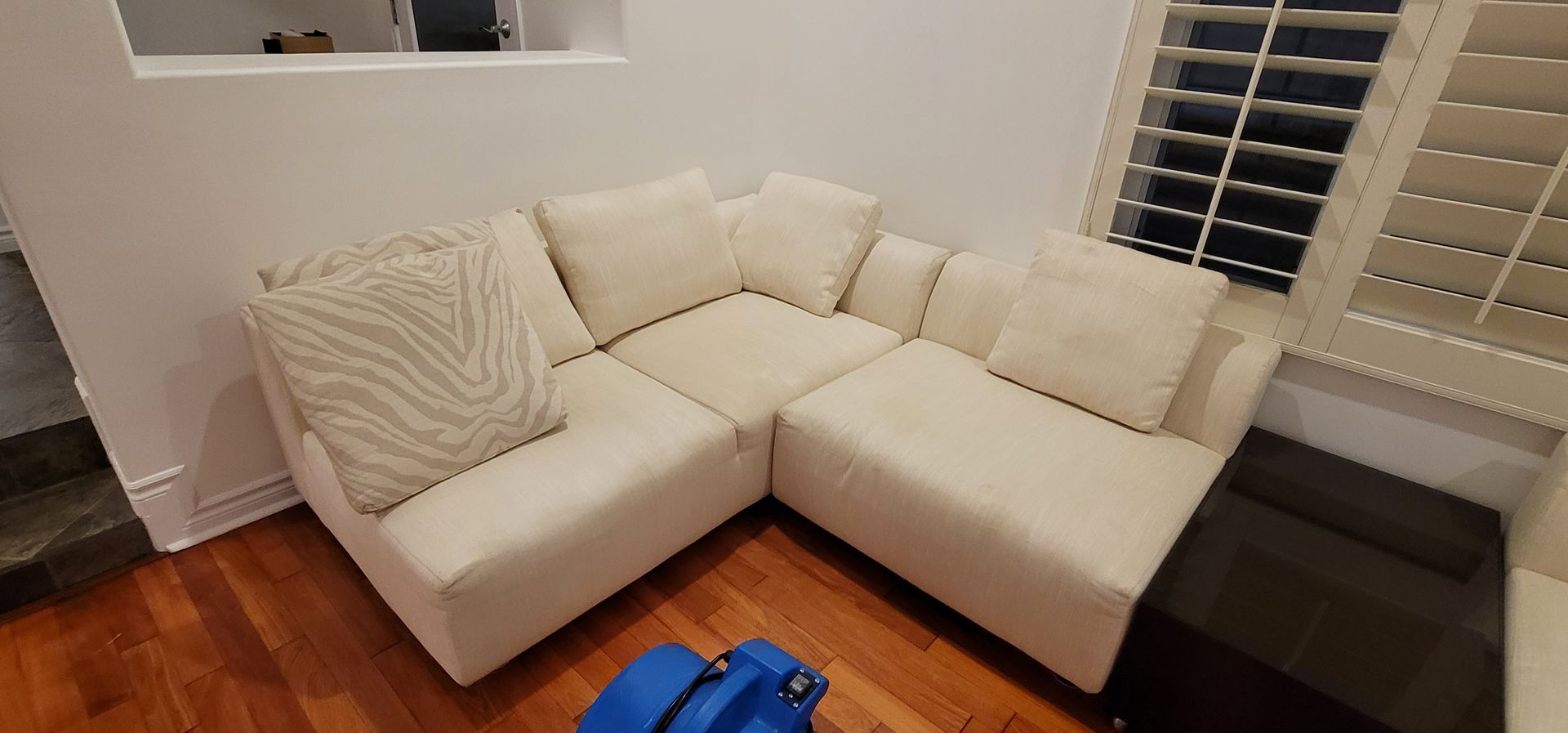 Cream sectional sofa in a corner by windows with blinds on a hardwood floor.