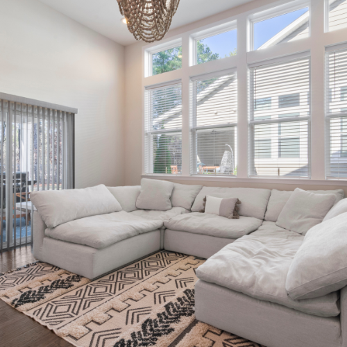 Bright living room with a large white sectional sofa, patterned rug, and tall windows with blinds.