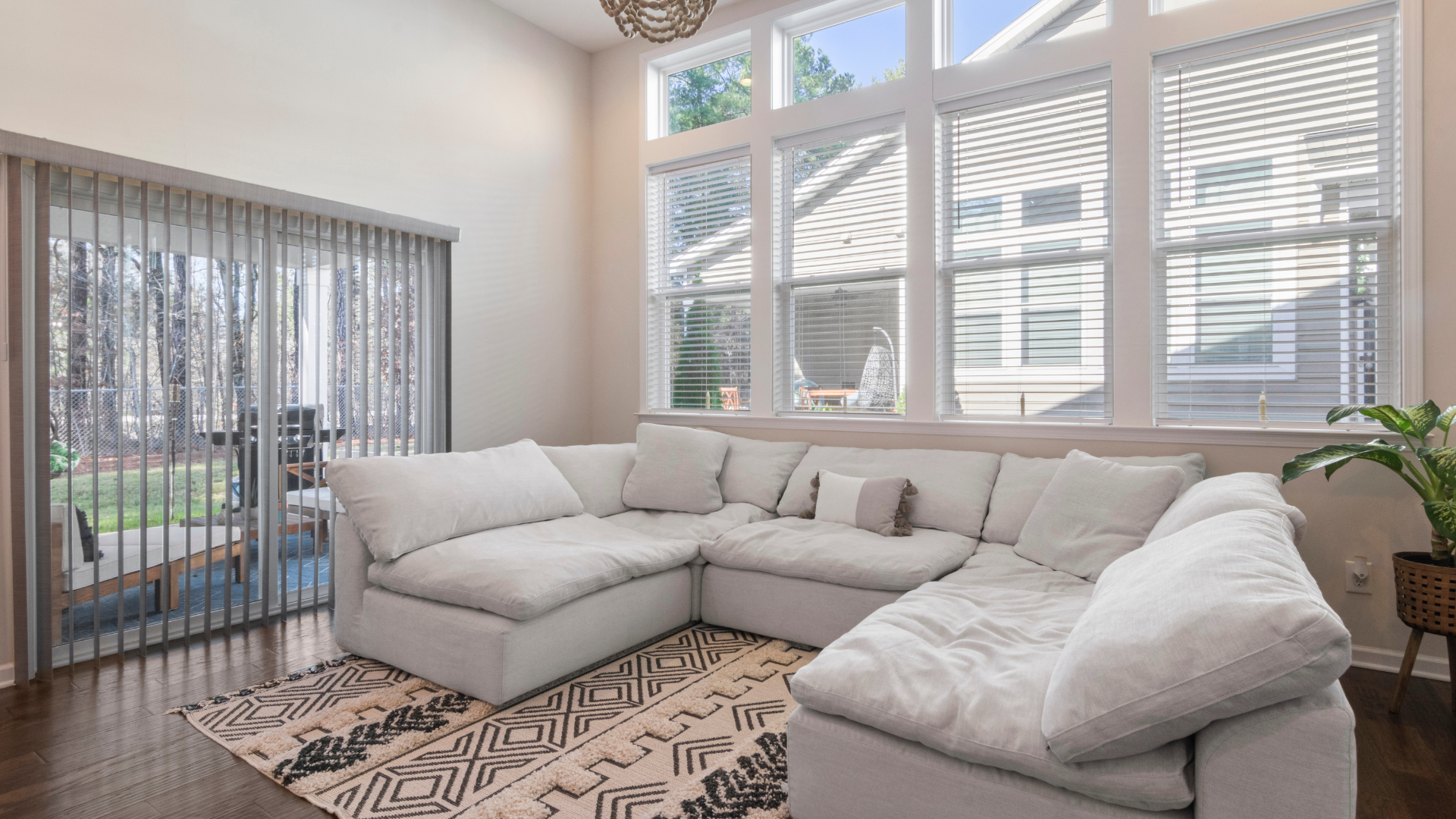 Bright living room with a large gray sectional, patterned rug, and floor-to-ceiling windows with blinds.