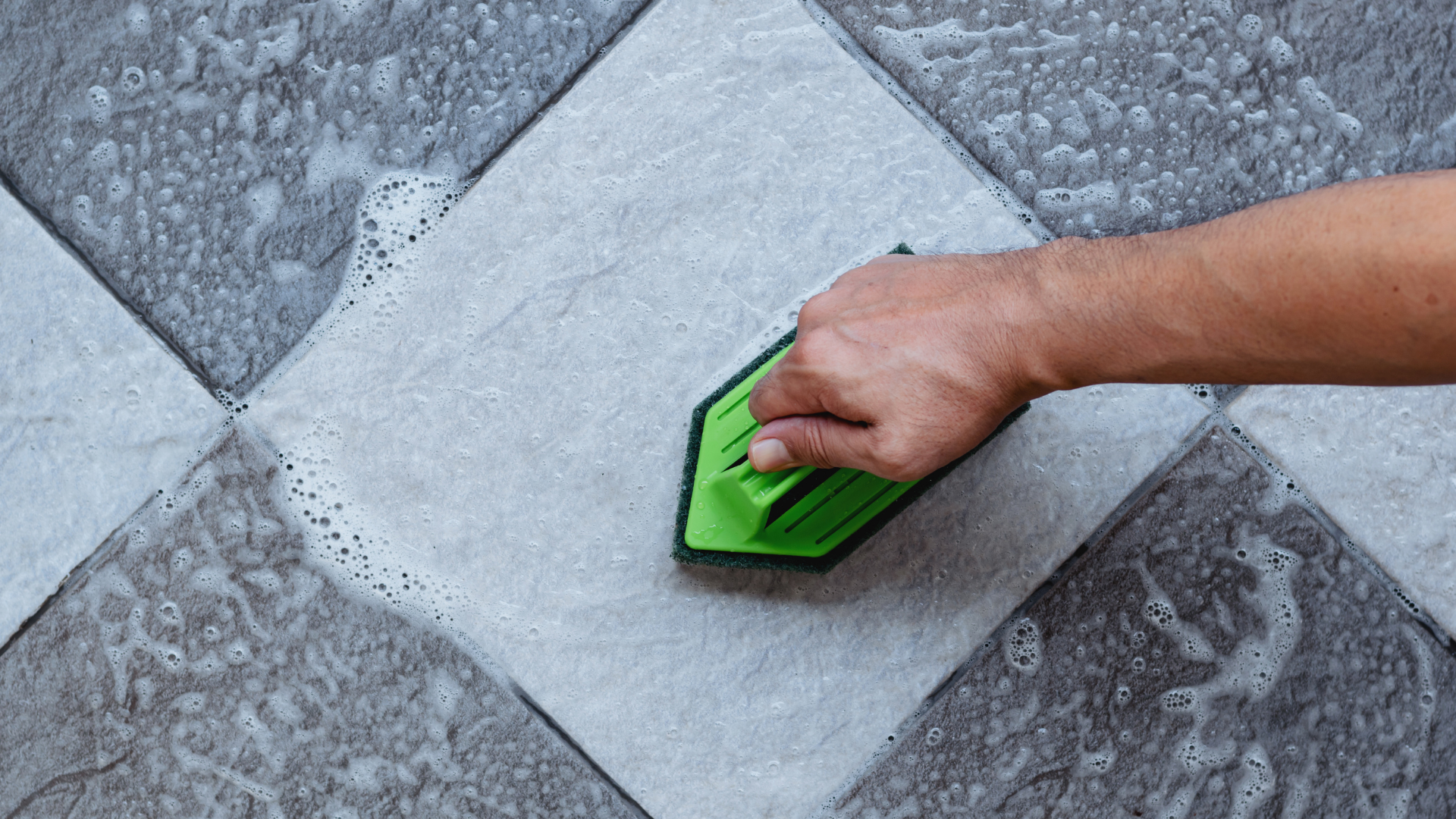 Hand wiping a gray tiled floor with a green cleaning pad