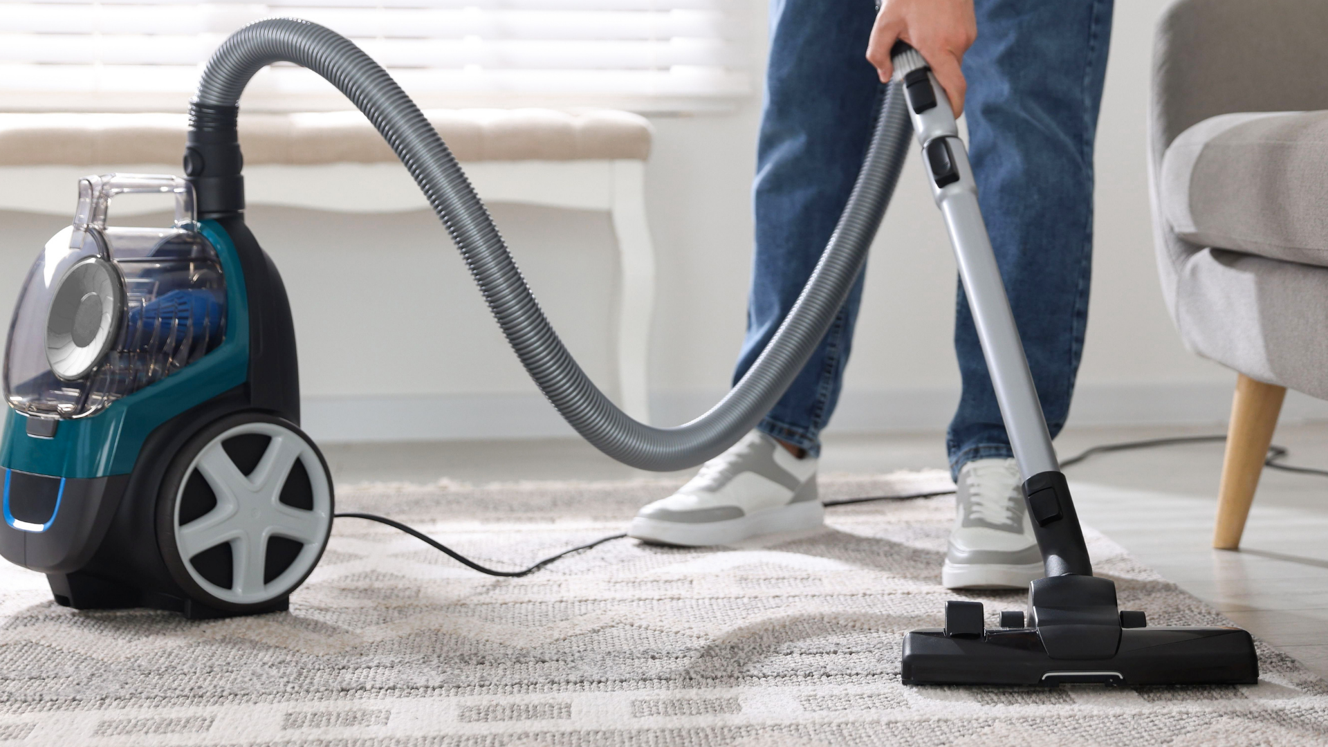 Person vacuuming a light rug with a blue canister vacuum in a bright living room