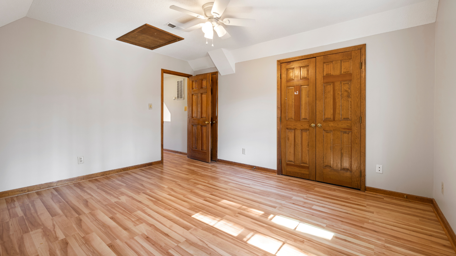 Empty bedroom with hardwood floors, white walls, ceiling fan, and wooden closet doors in bright sunlight.