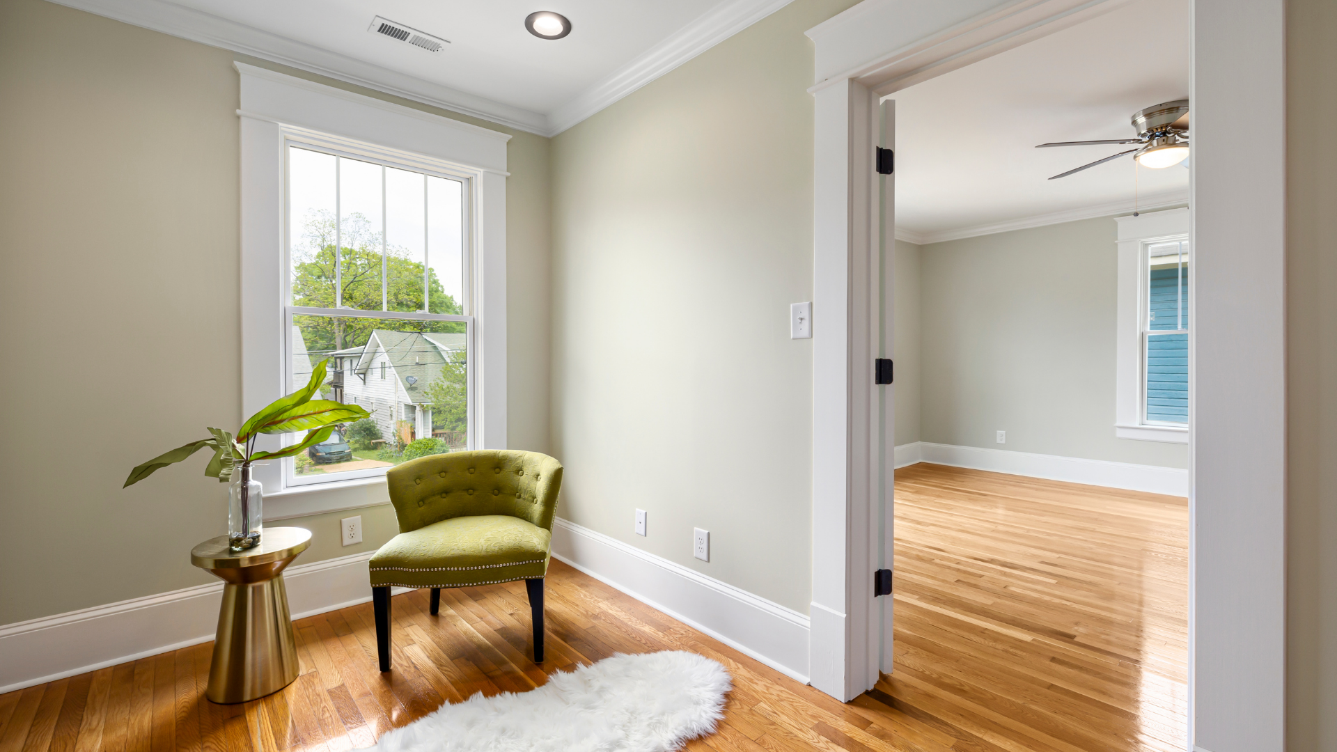 Bright empty room with large window, green chair, side table, and hardwood floors opening to another room
