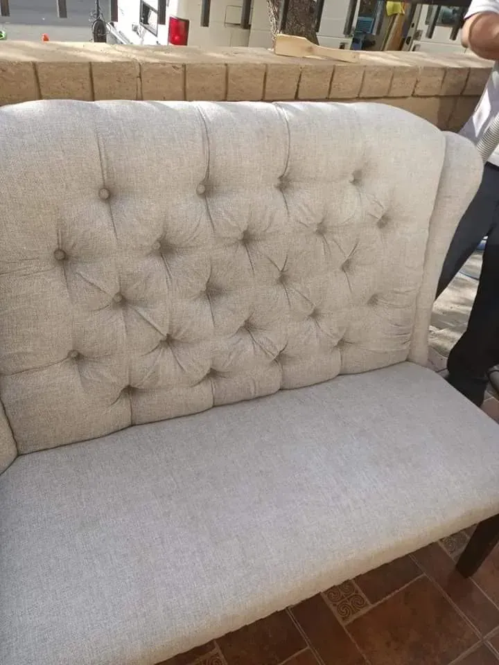 Light gray tufted upholstered loveseat with a high buttoned back and cushioned seat outdoors.
