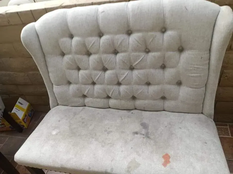 Light gray tufted loveseat with worn seat, against a brick wall.