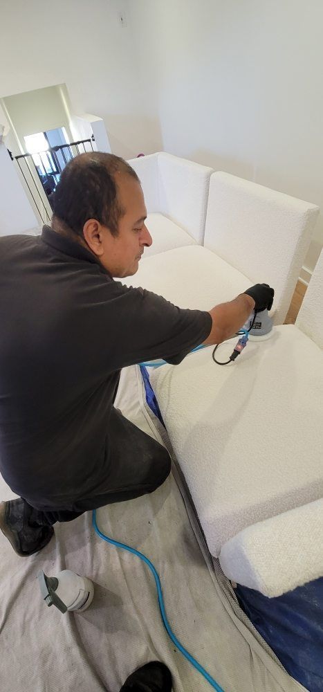 Person vacuuming a white sofa in a bright living room, with a blue cord on the floor