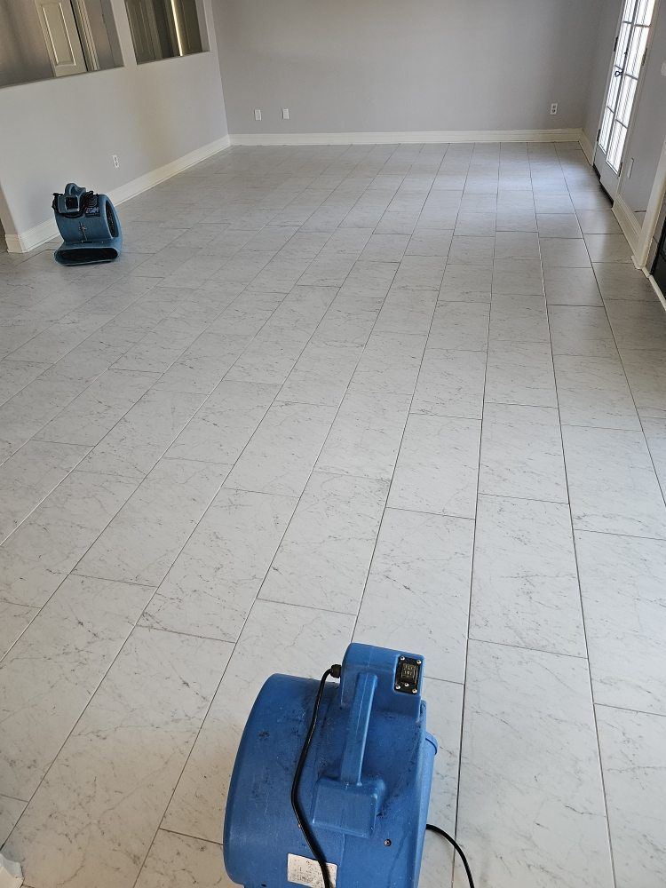 Vacant tiled room with beige walls, blue floor buffer in foreground, and another cleaner near the back.