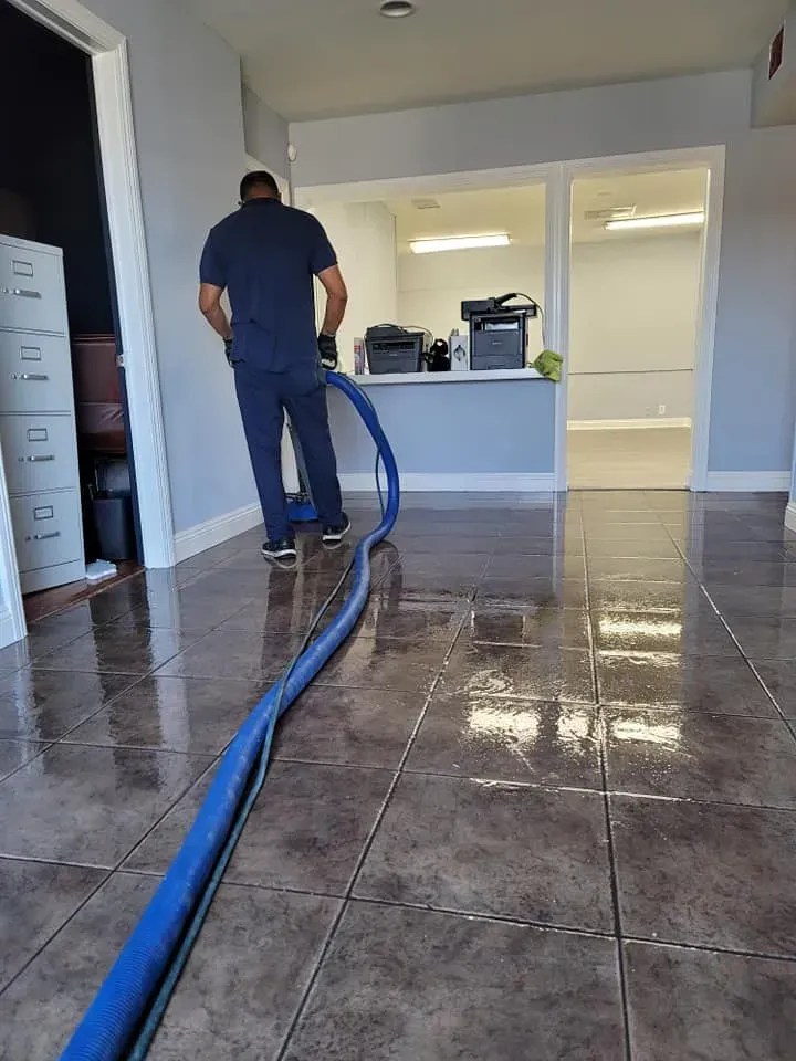 Person cleaning a tiled floor with a blue hose in a bright indoor room