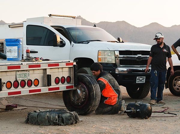 Worker changing a tire on a white service truck in a desert setting, with another person standing nearby