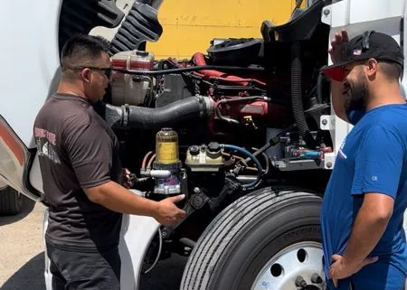 Two men inspecting a truck engine, outdoors. One points, the other looks on.