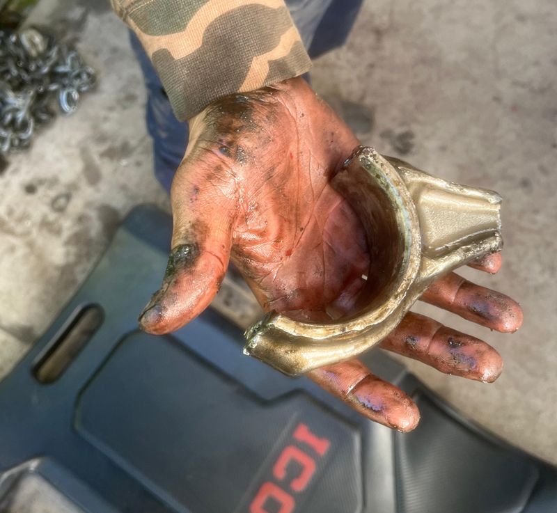 Hand holding a broken metal automotive part, covered in oil, against a camo sleeve and black mat.