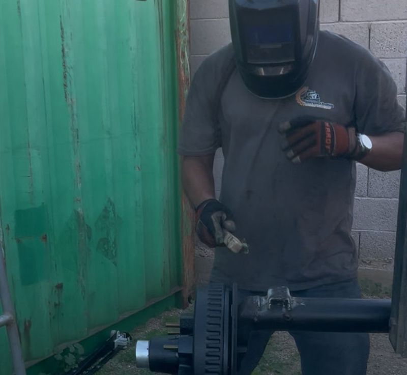 Welder in a mask, welding on a trailer axle outdoors, near a green container.