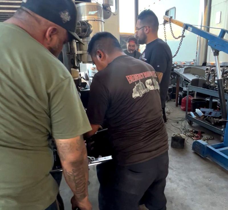 Three men working on machinery in a workshop. One wears a black t-shirt, another a green one, and a third watches.