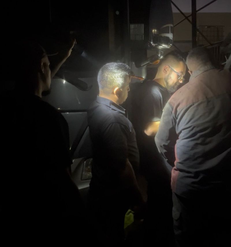 Group of people standing near a vehicle at night, illuminated by lights. Some are looking down.