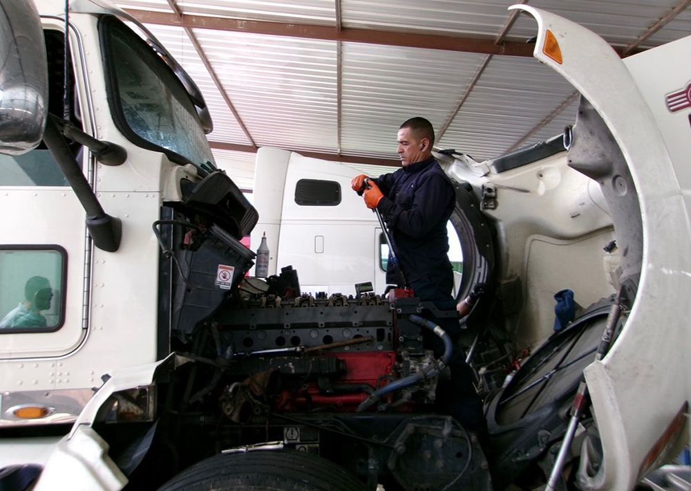 Mechanic working on the engine of a semi-truck inside a garage; truck hood open.