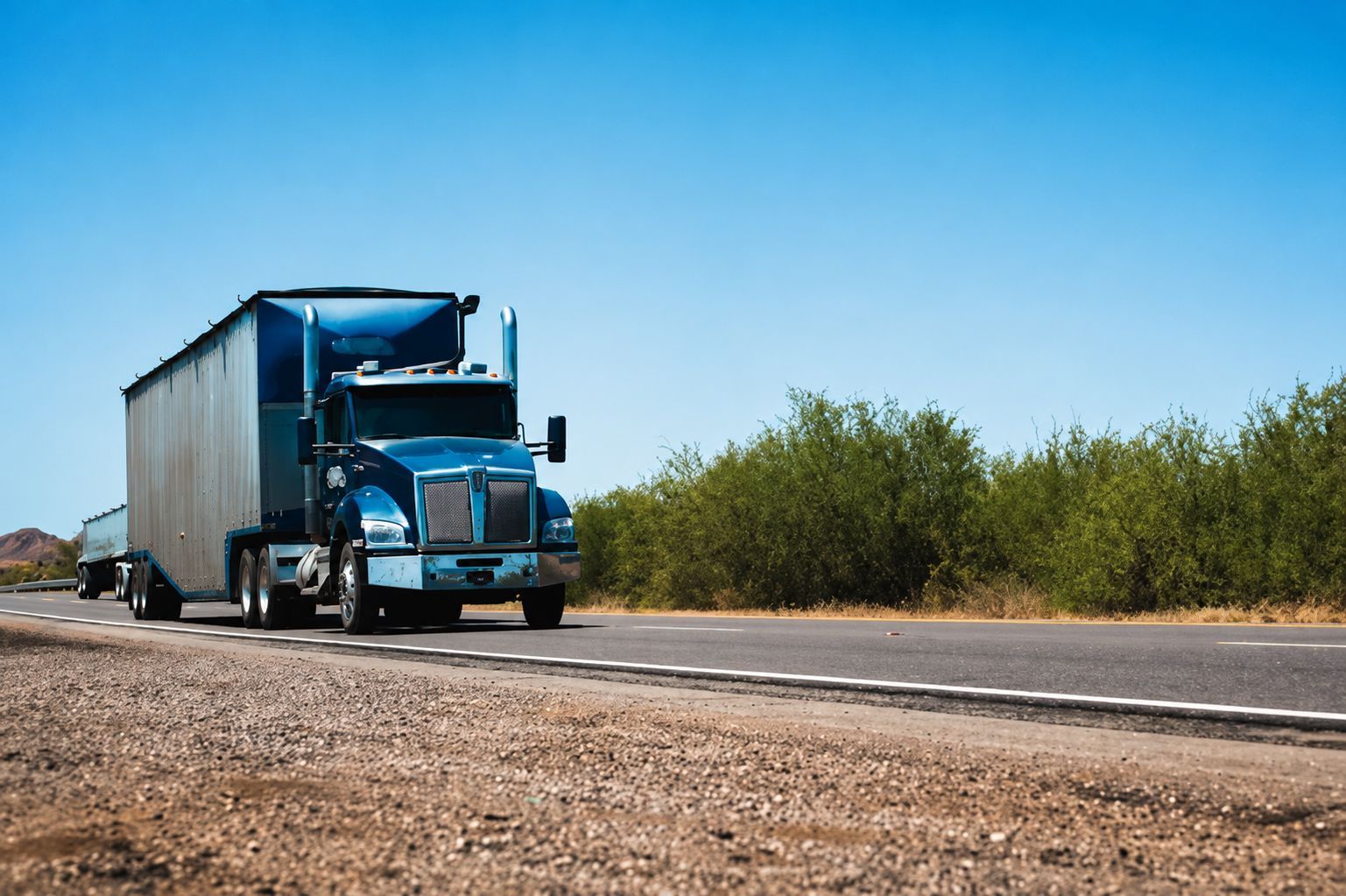 Blue semi-truck driving on a rural highway beside desert shrubs under a clear sky