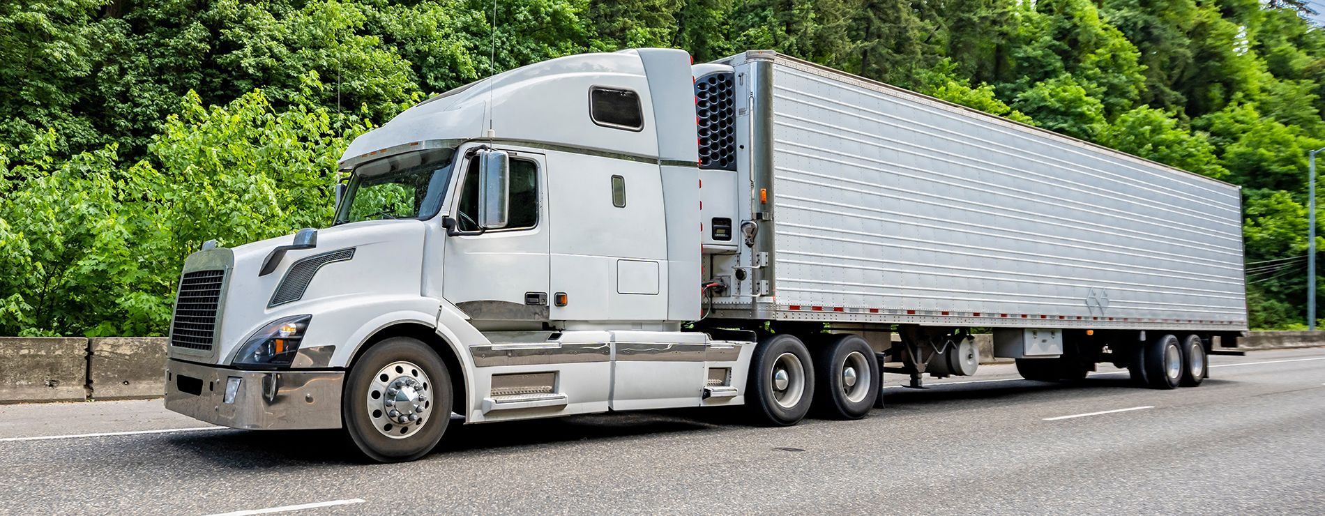 White semi-truck with trailer driving on a highway, with green foliage in the background.