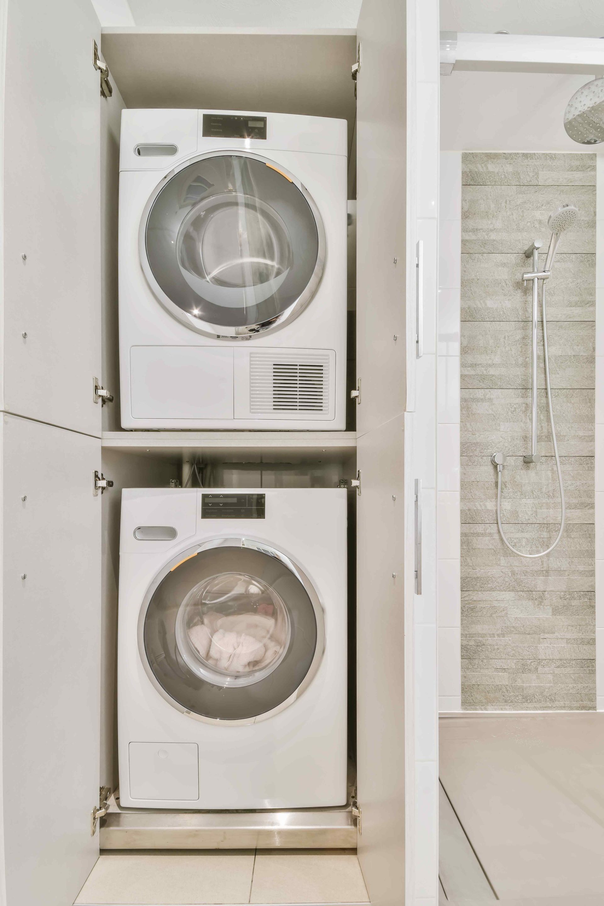 Stacked white washer and dryer units inside a cabinet, next to a shower.