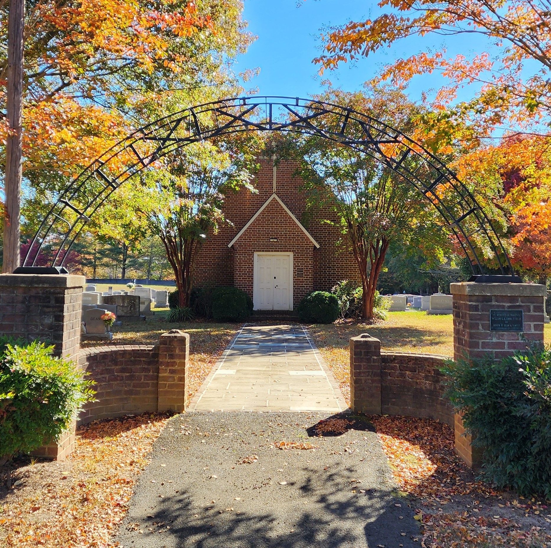 Brick chapel under a metal archway, framed by colorful fall trees. Entrance path, cemetery in background.