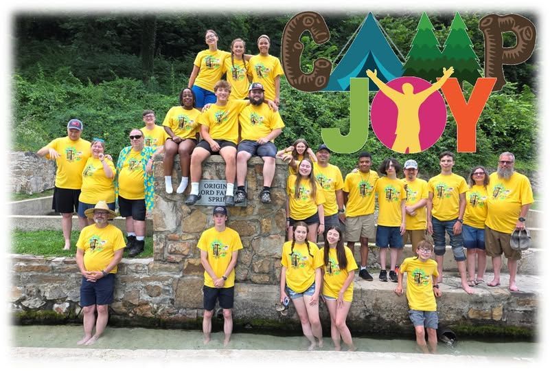 Camp Joy group photo. People in yellow shirts standing/sitting by water. CAMP Joy logo with tent and person.