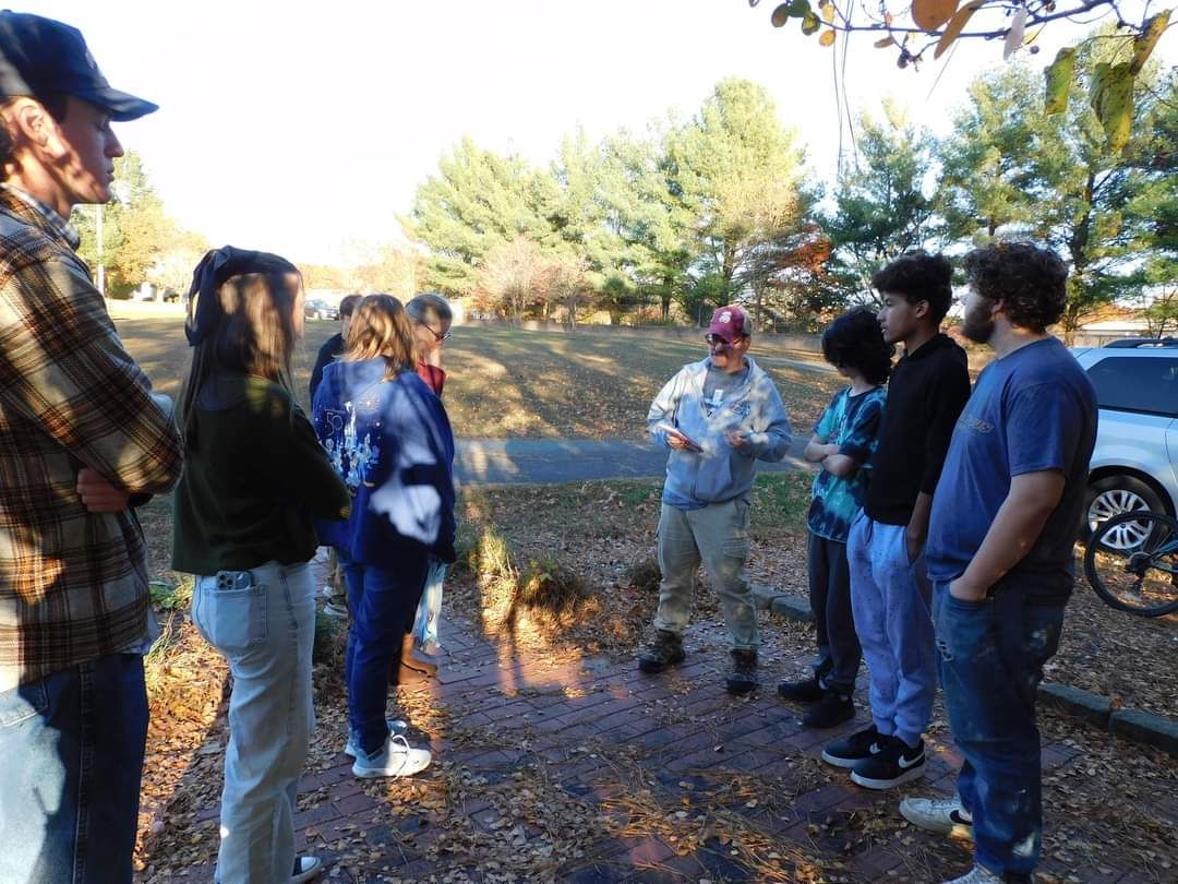 A group of people stands outside, listening to a person in the center. The setting is a yard with trees.
