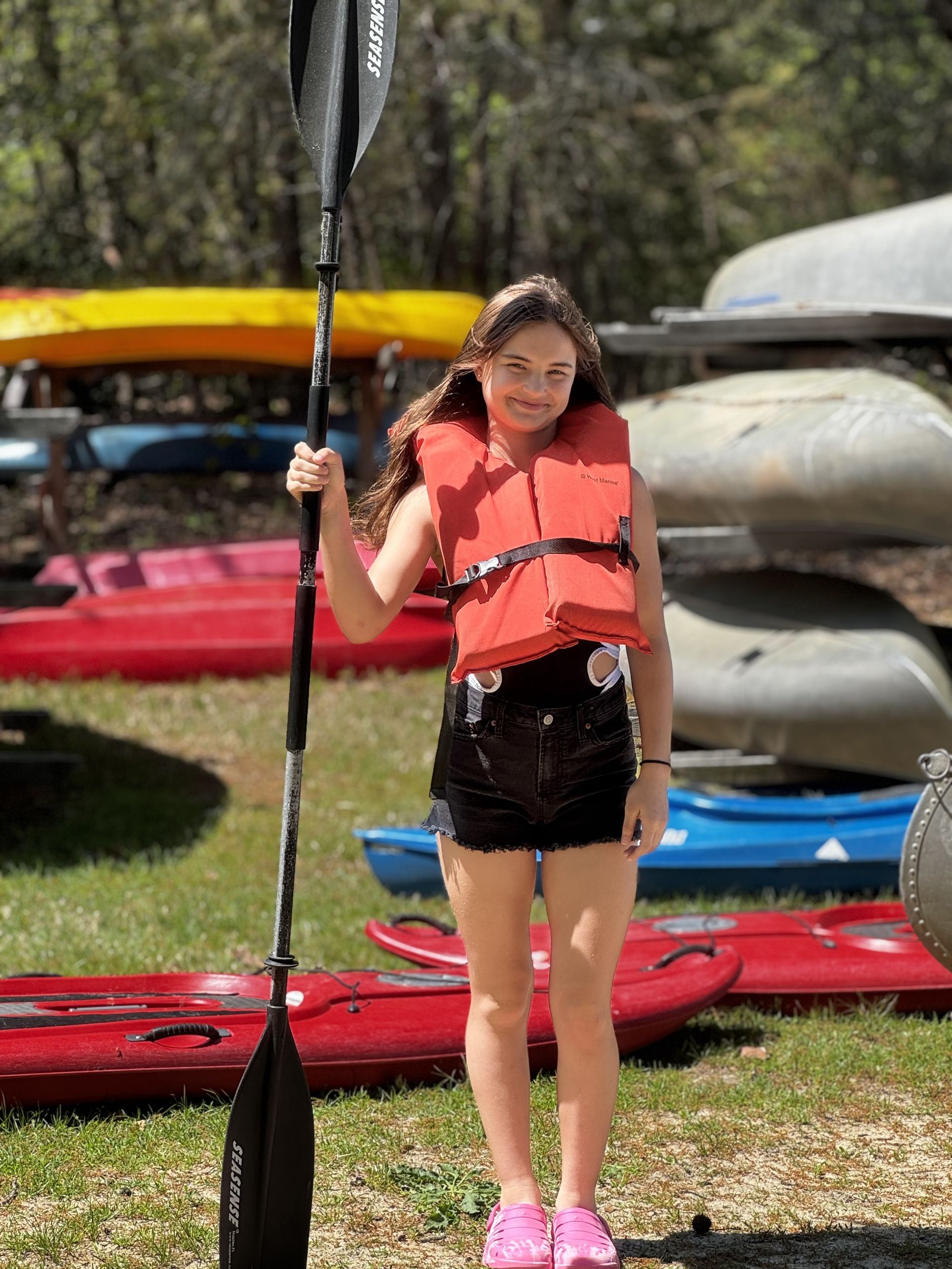 A youth in a orange life jacket holding a paddle beside colorful kayaks on a grassy shore
