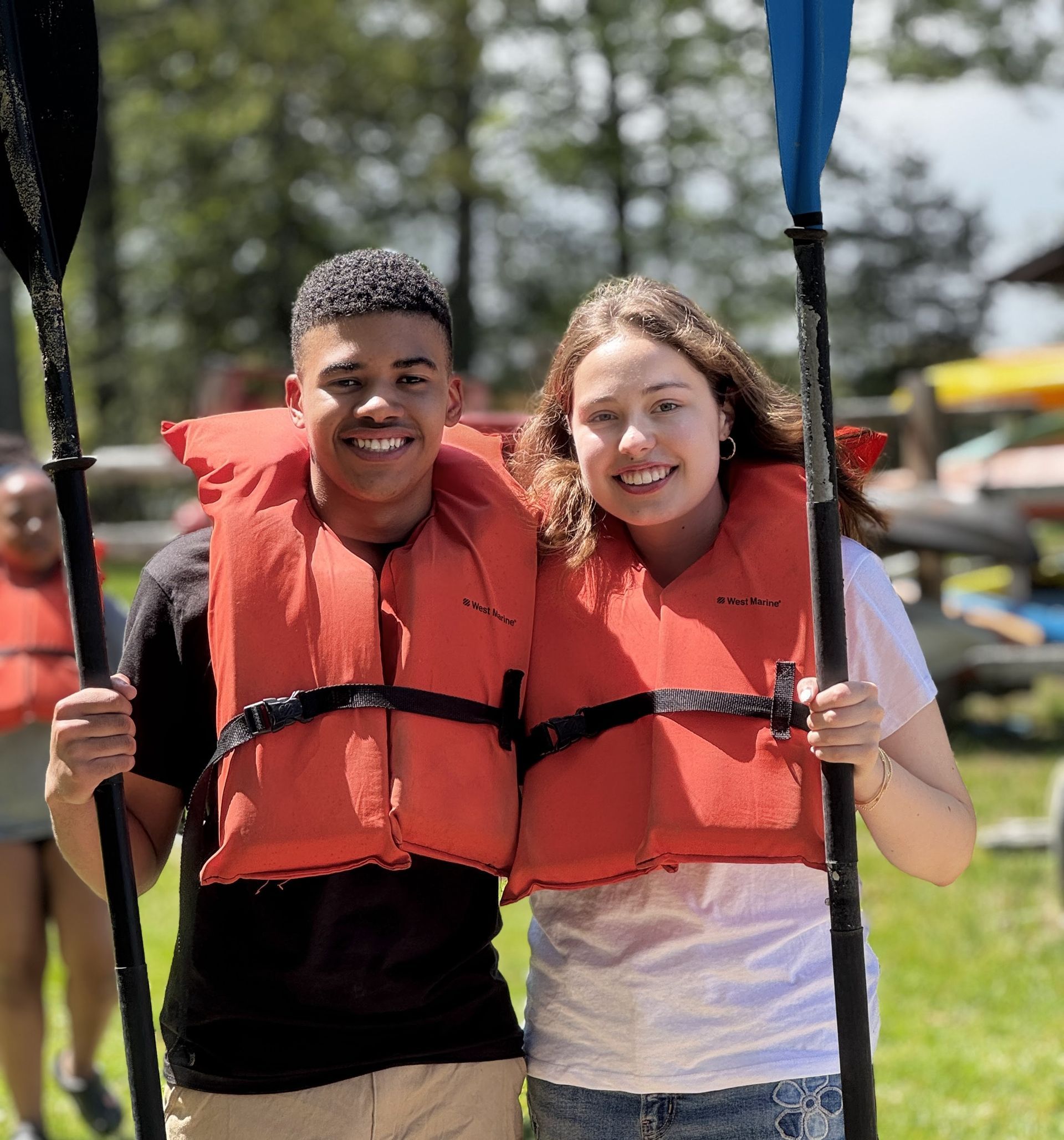 Two youth smiling in orange life jackets, holding paddles outdoors near the water.