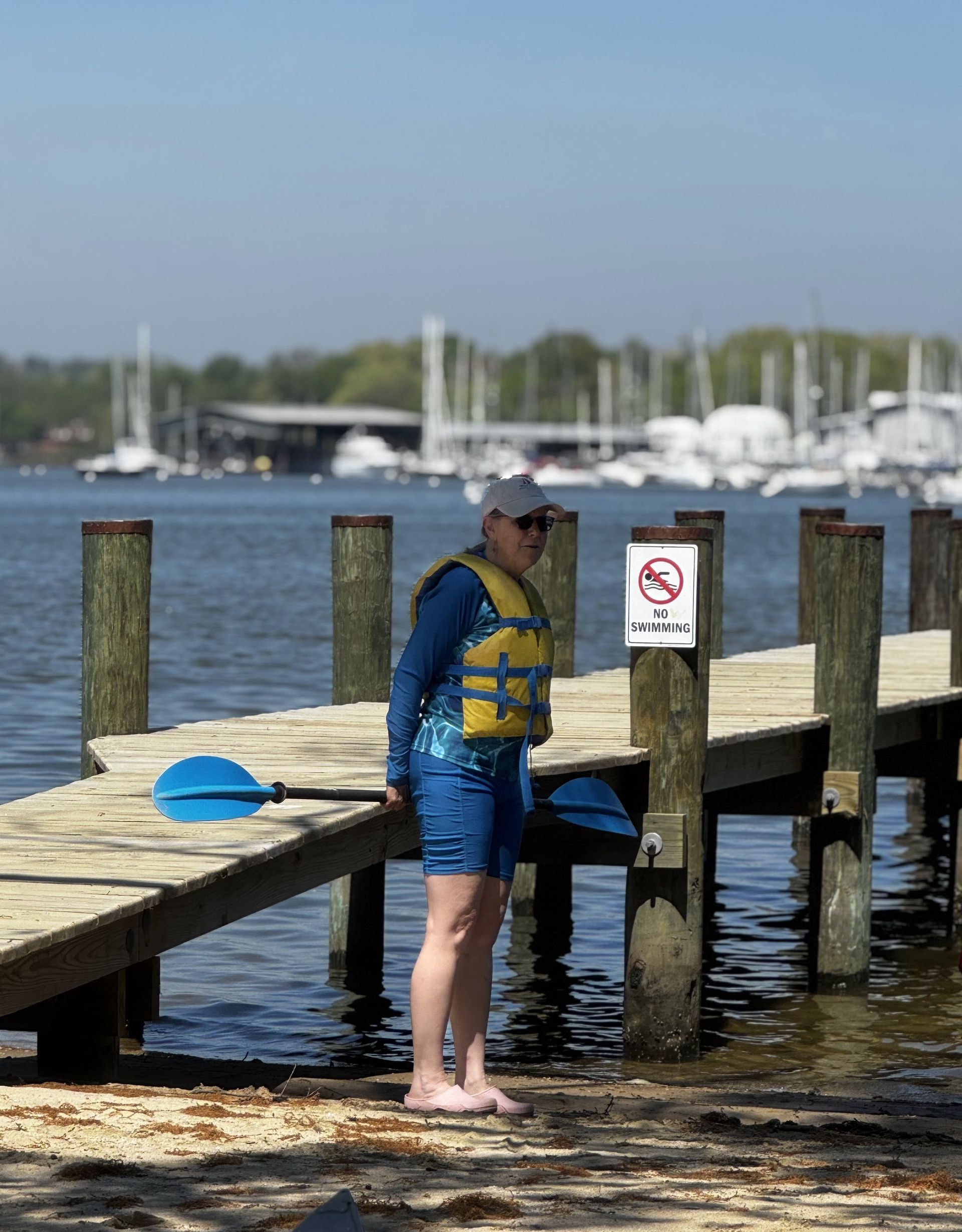 A youth leader in blue life jacket standing on a dock beside a lake, with sailboats in the background.