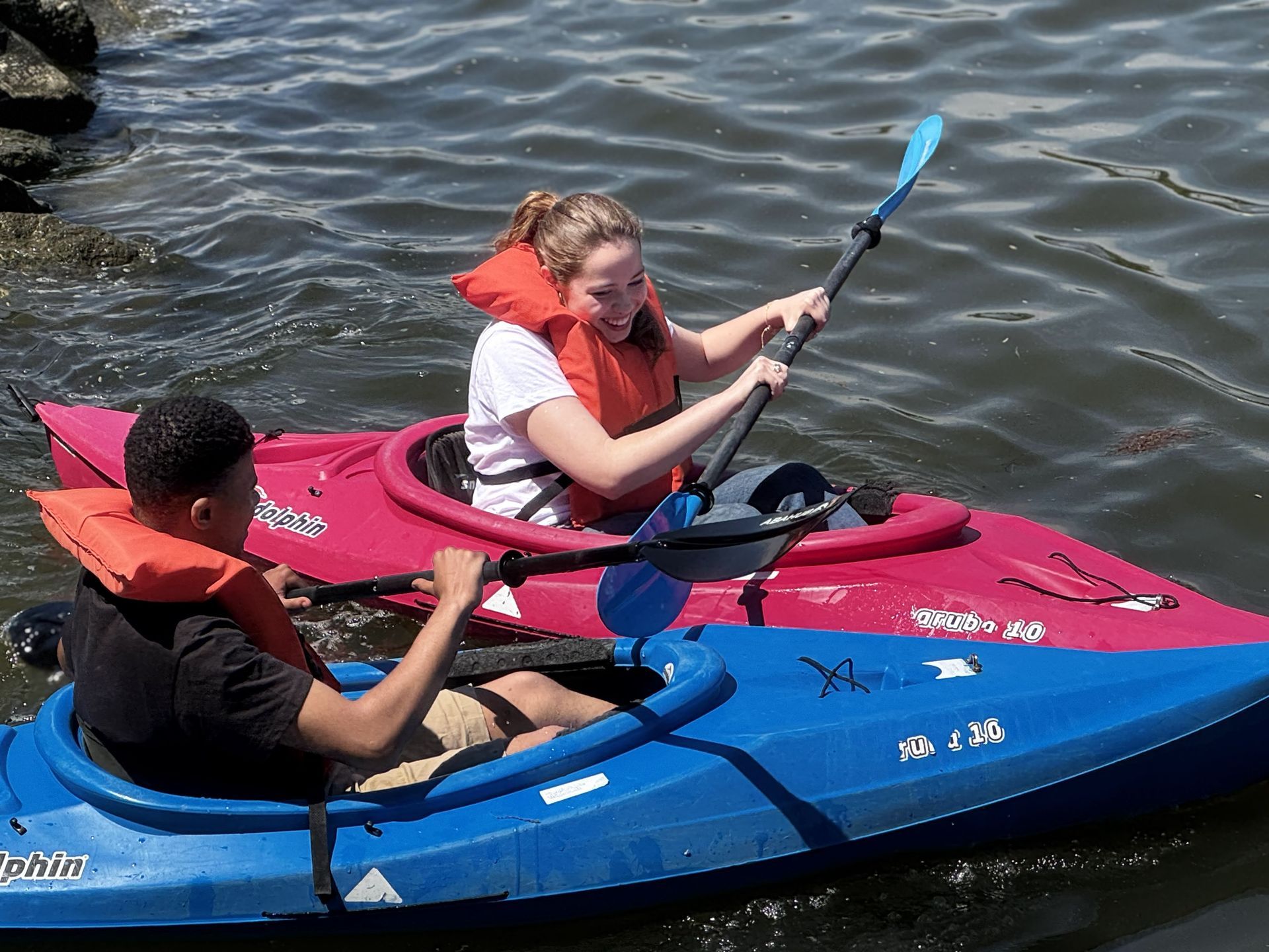 Two people kayaking on rippling water, one in a blue kayak and one in a pink kayak, paddling side by side