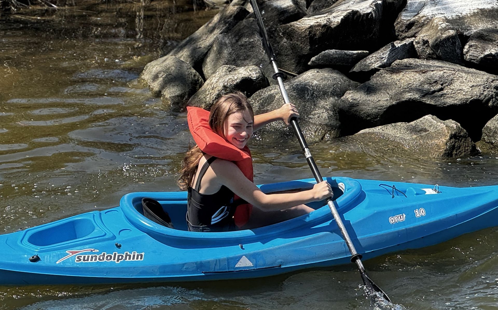 Person kayaking in a blue kayak near rocky shoreline, paddling on calm water.