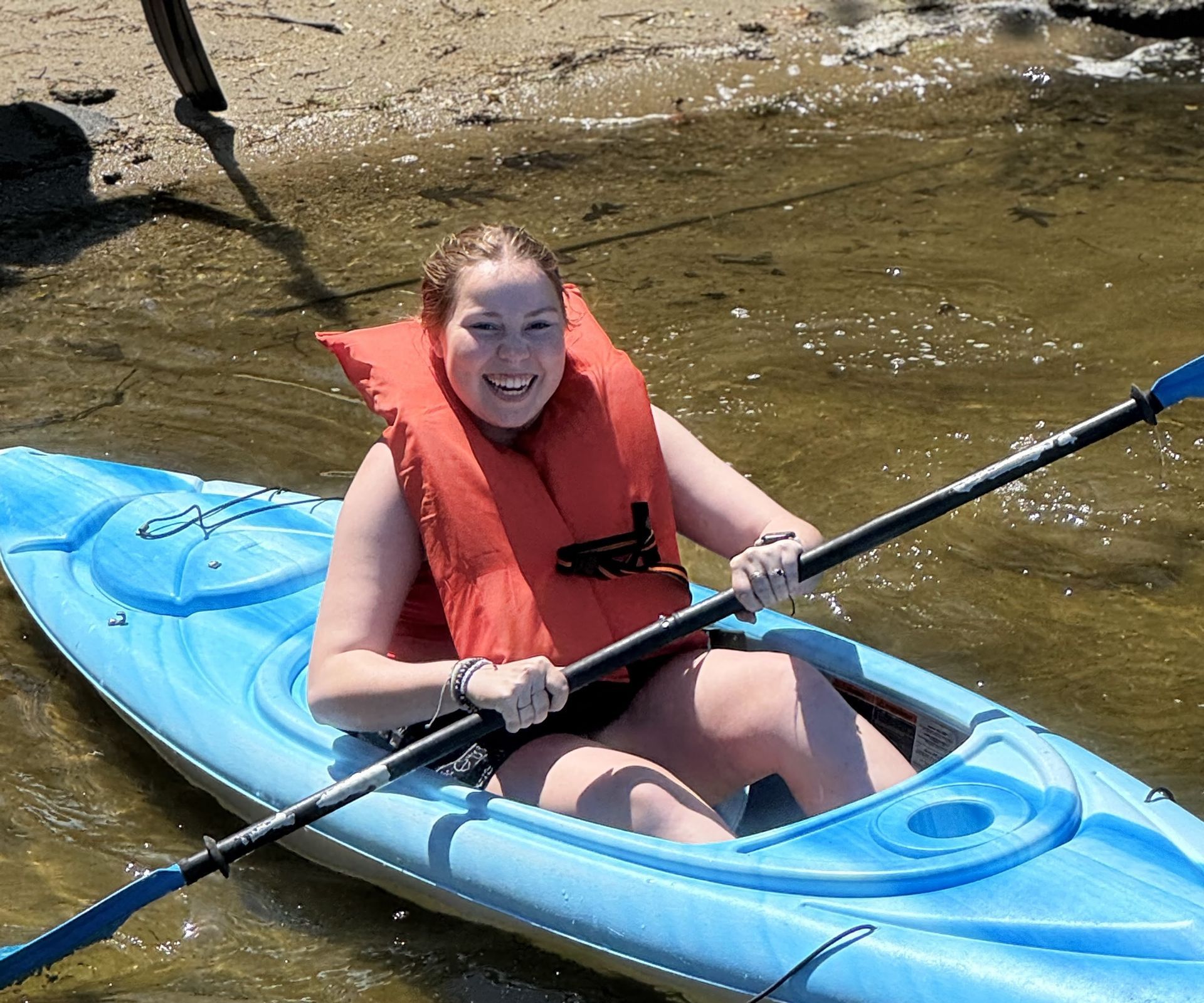 A youth in a blue kayak on shallow water, smiling and paddling with a red life jacket.