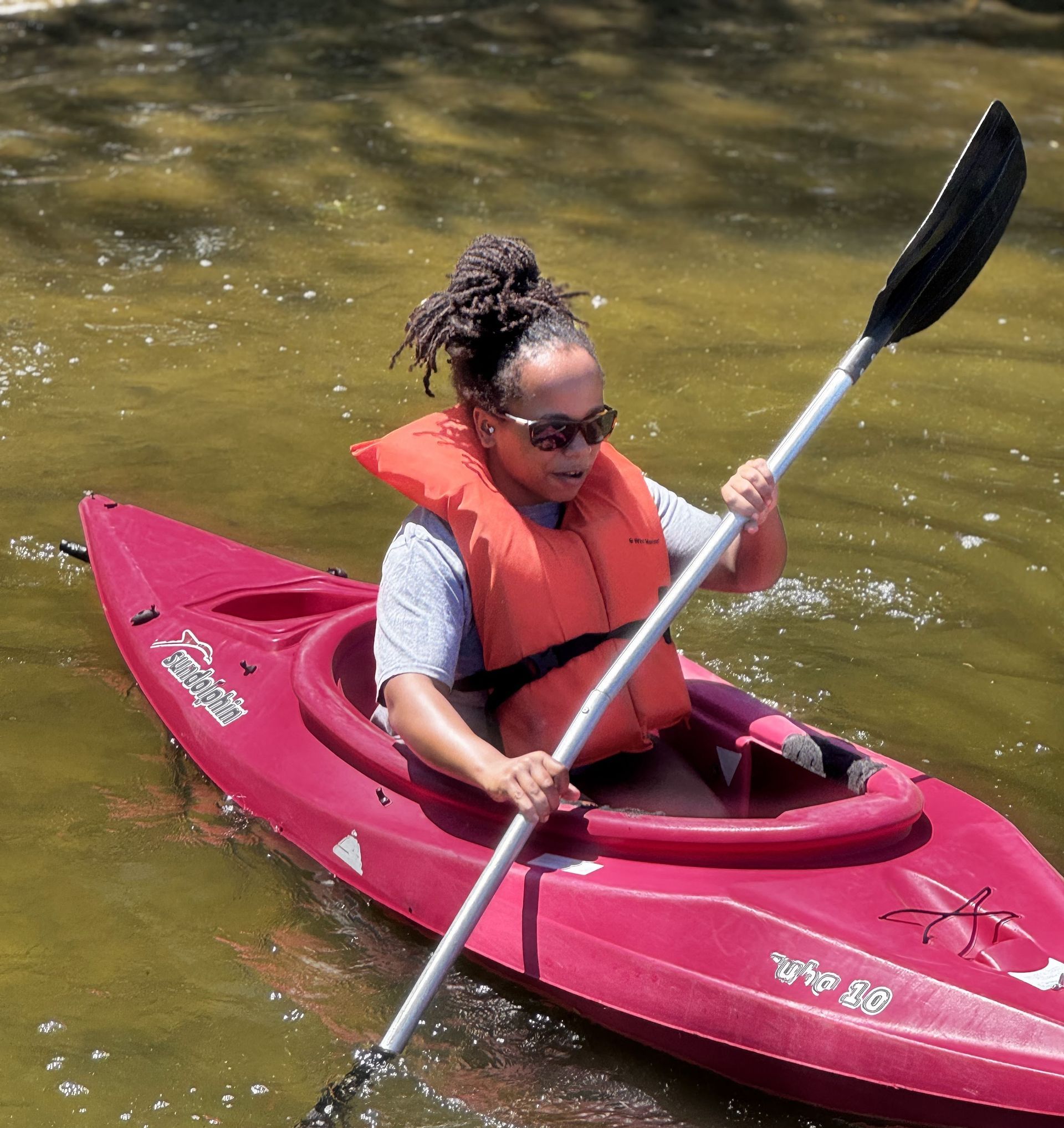 A youth paddling a pink kayak on calm water, wearing an orange life vest and sunglasses