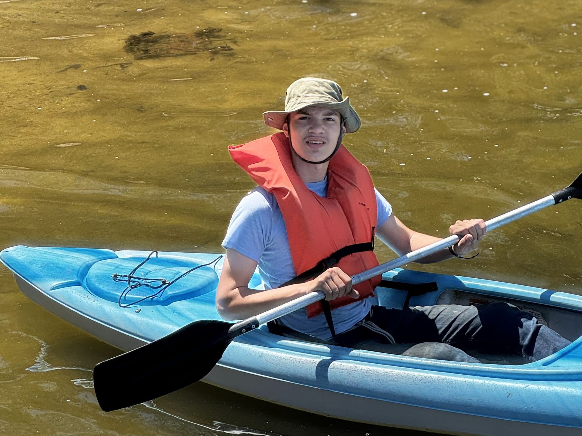 Person in a blue kayak wearing a orange life vest, paddling on calm water under sunlight