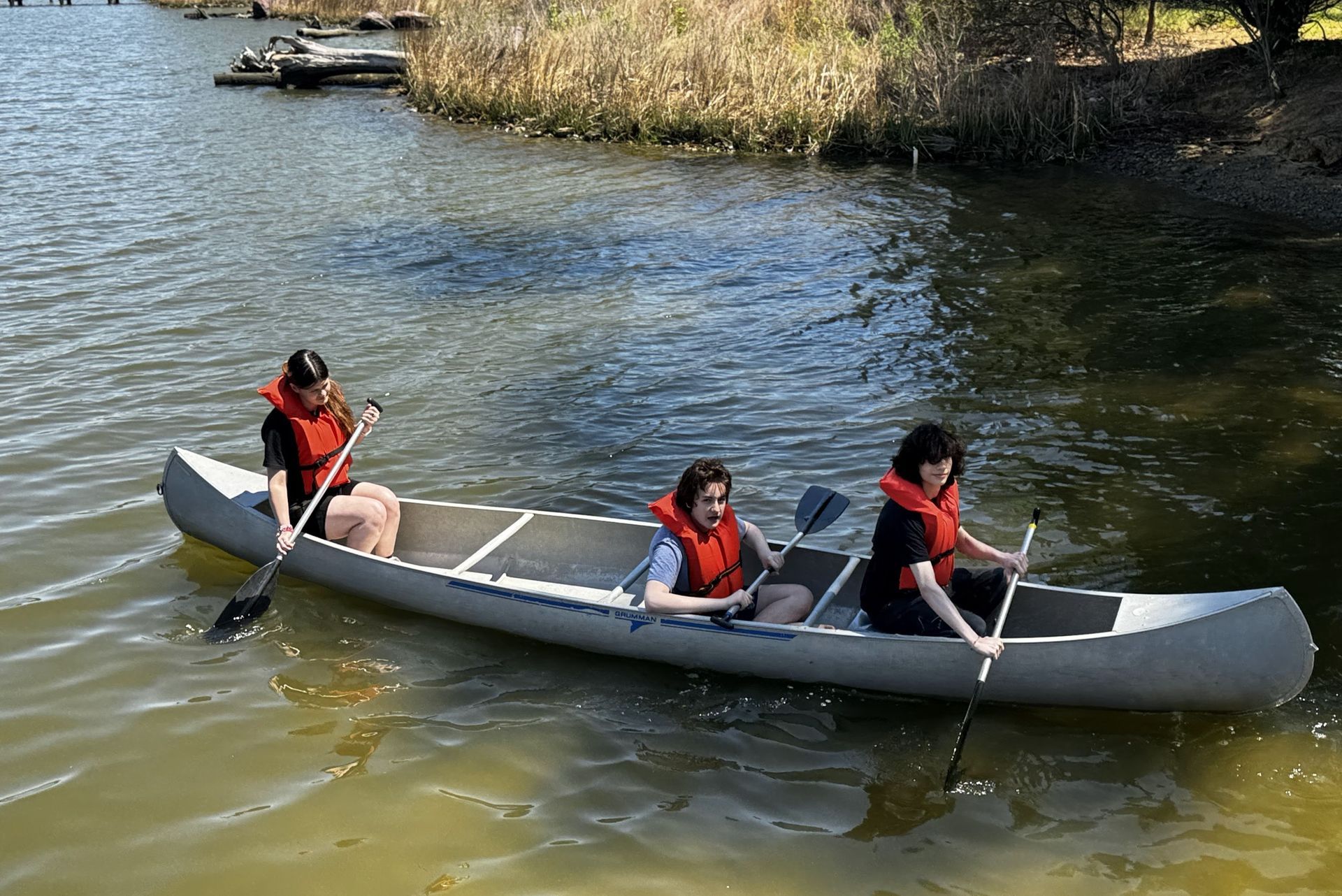 Three people in life jackets paddling a canoe on a calm river near reeds