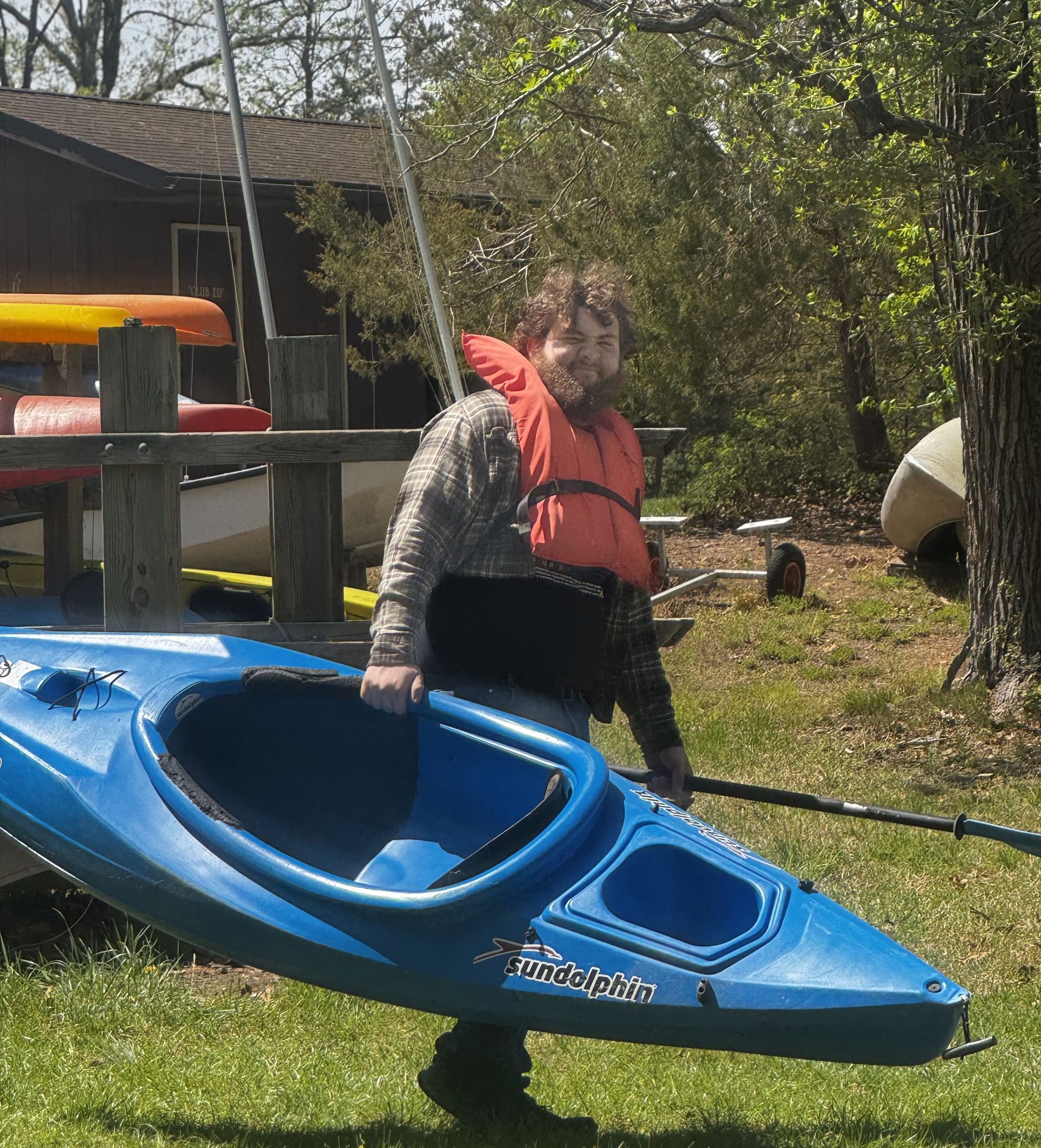 Person in orange life jacket standing beside a blue kayak outdoors near a house and trees