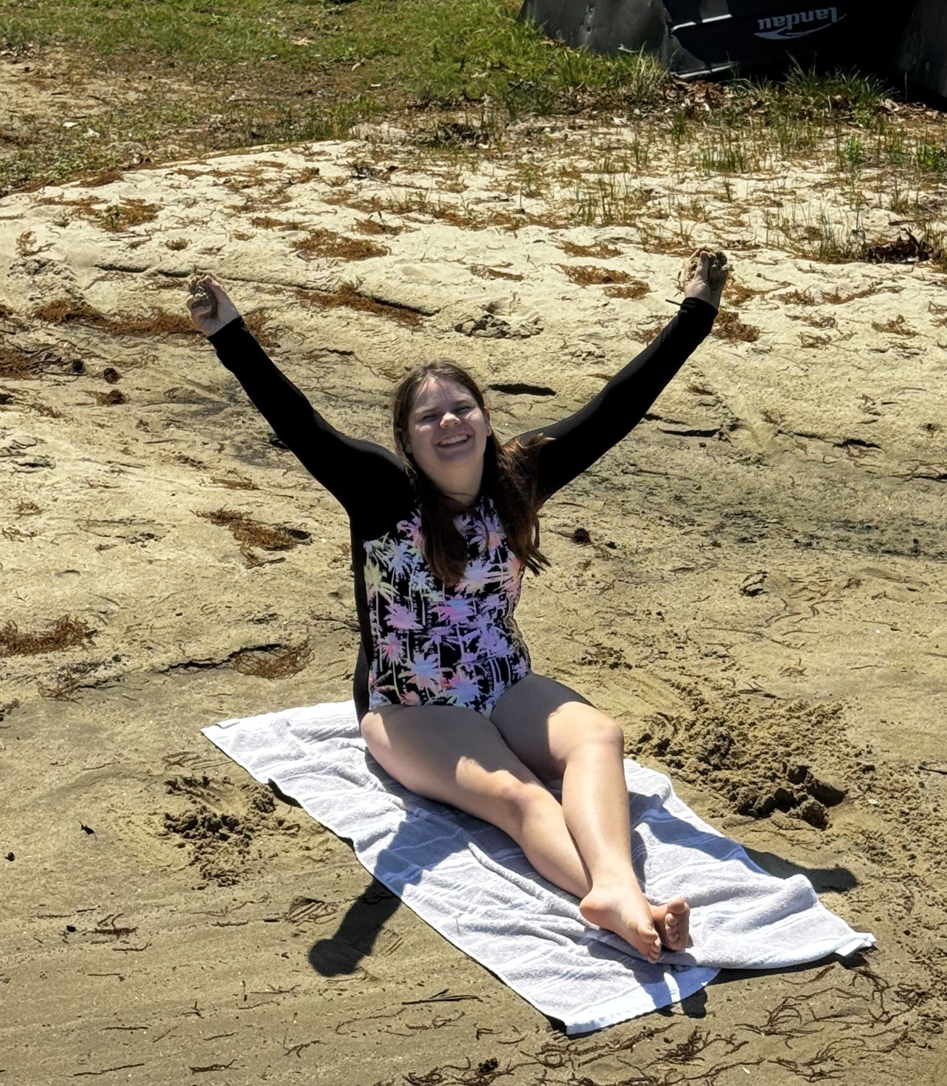 A youth sitting on a towel at the beach, smiling with arms raised in the air.