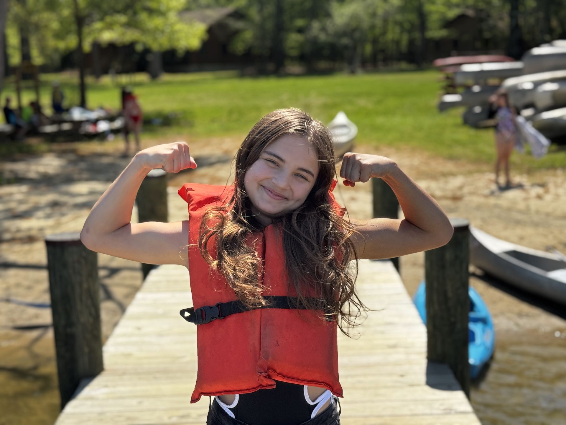 A youth in a red life jacket flexing arms on a dock by the water, smiling outdoors.