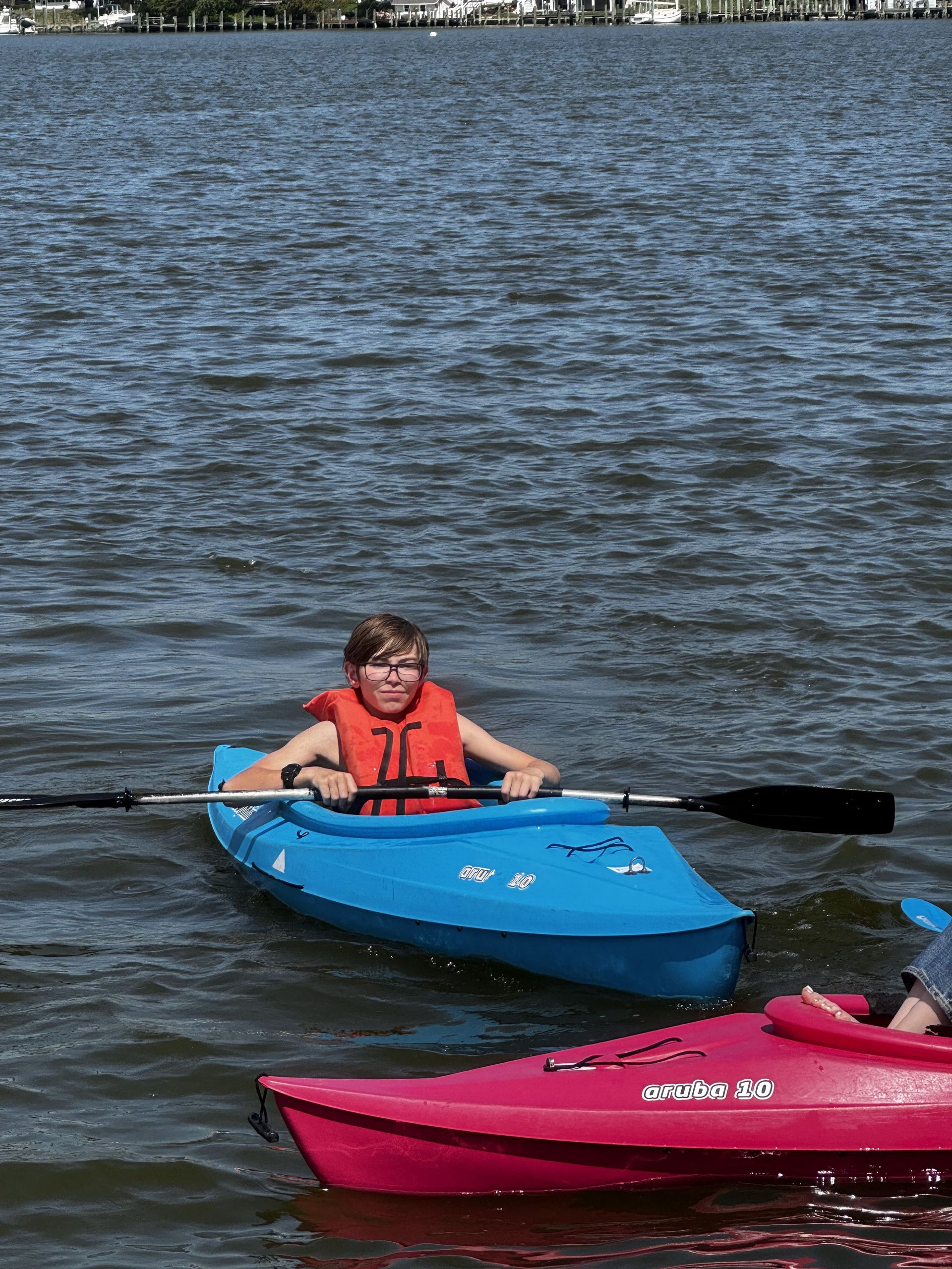 Two kayakers on the water, one in a blue kayak wearing a orange life vest, another in a pink kayak nearby.