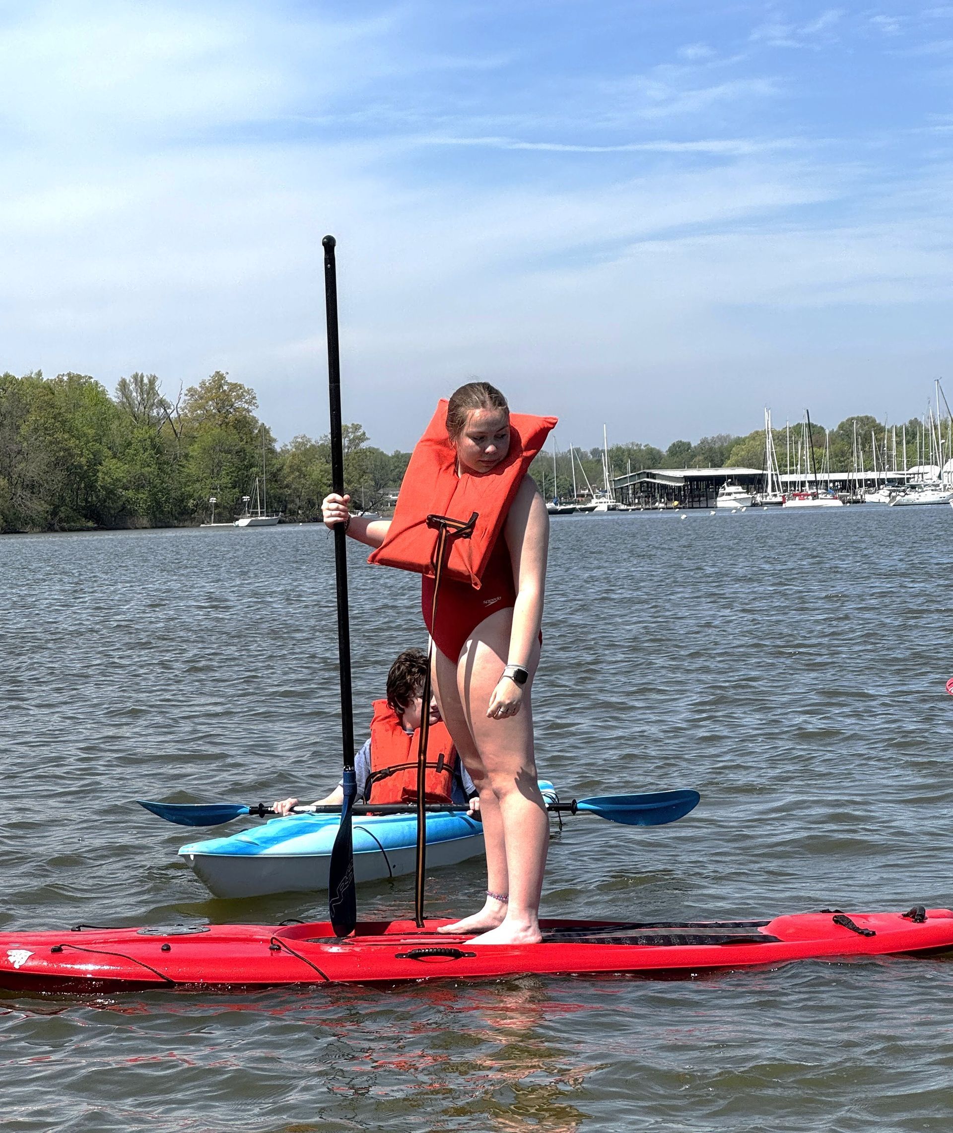 Person standing on a red paddleboard with a seated passenger on a lake, holding a paddle.