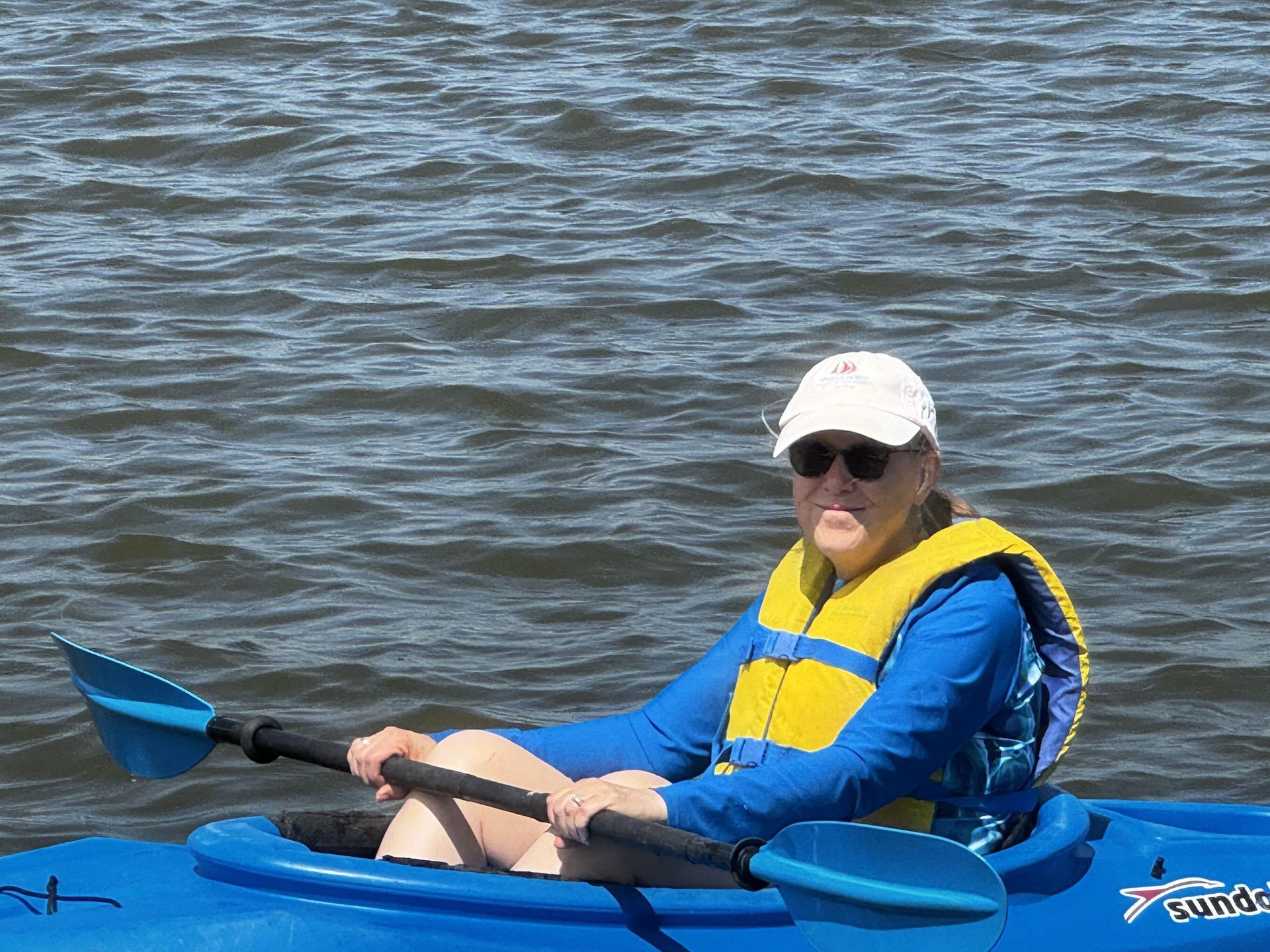 Person in a blue kayak on calm water, wearing a yellow life vest and white cap, holding a paddle