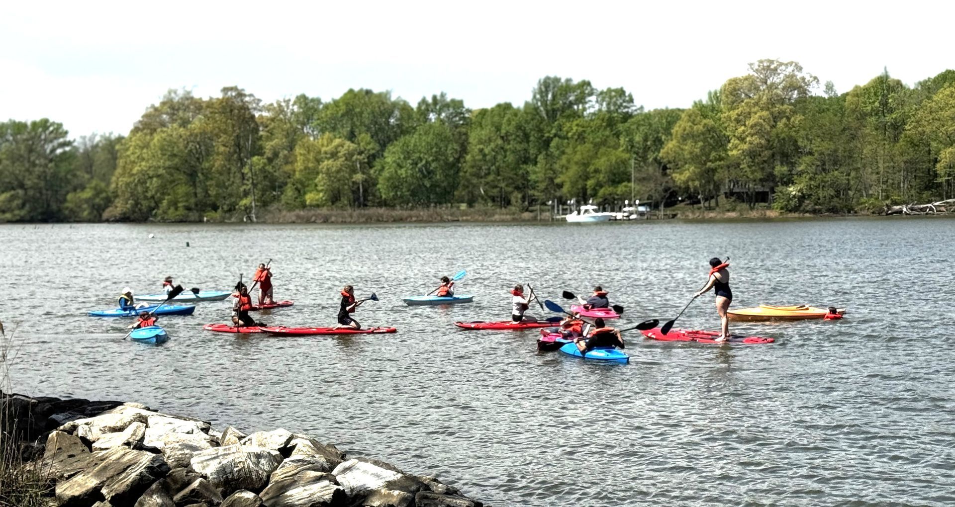 People kayaking on a lake near a rocky shore, with trees lining the far bank.
