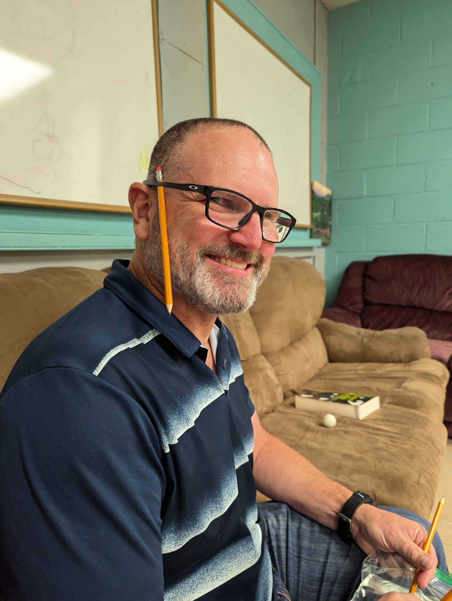 Man with a pencil behind his ear, wearing glasses, smiling; sitting on a couch.