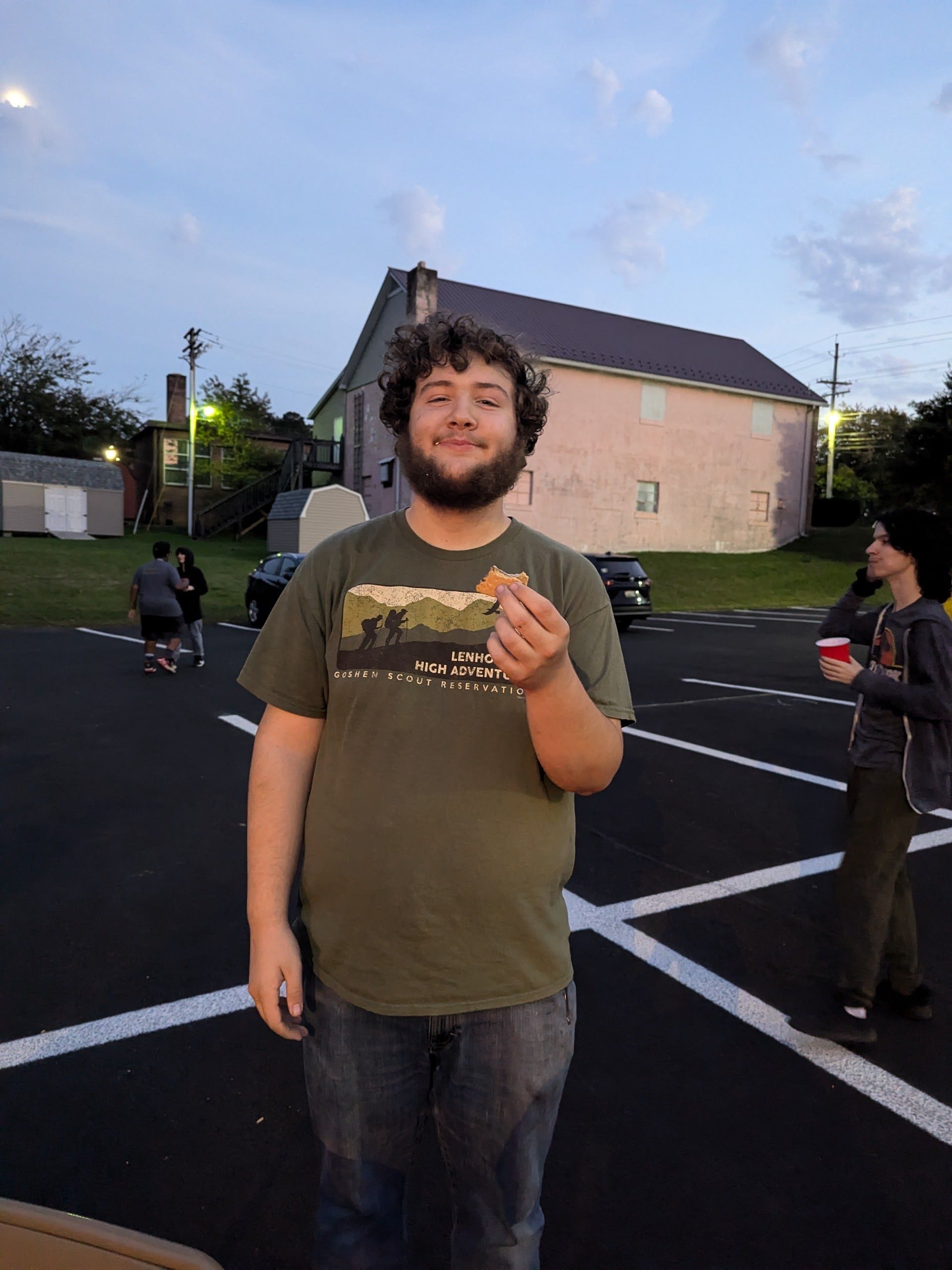 A man with curly hair, holding food, smiles in a parking lot. He wears a green shirt and jeans.